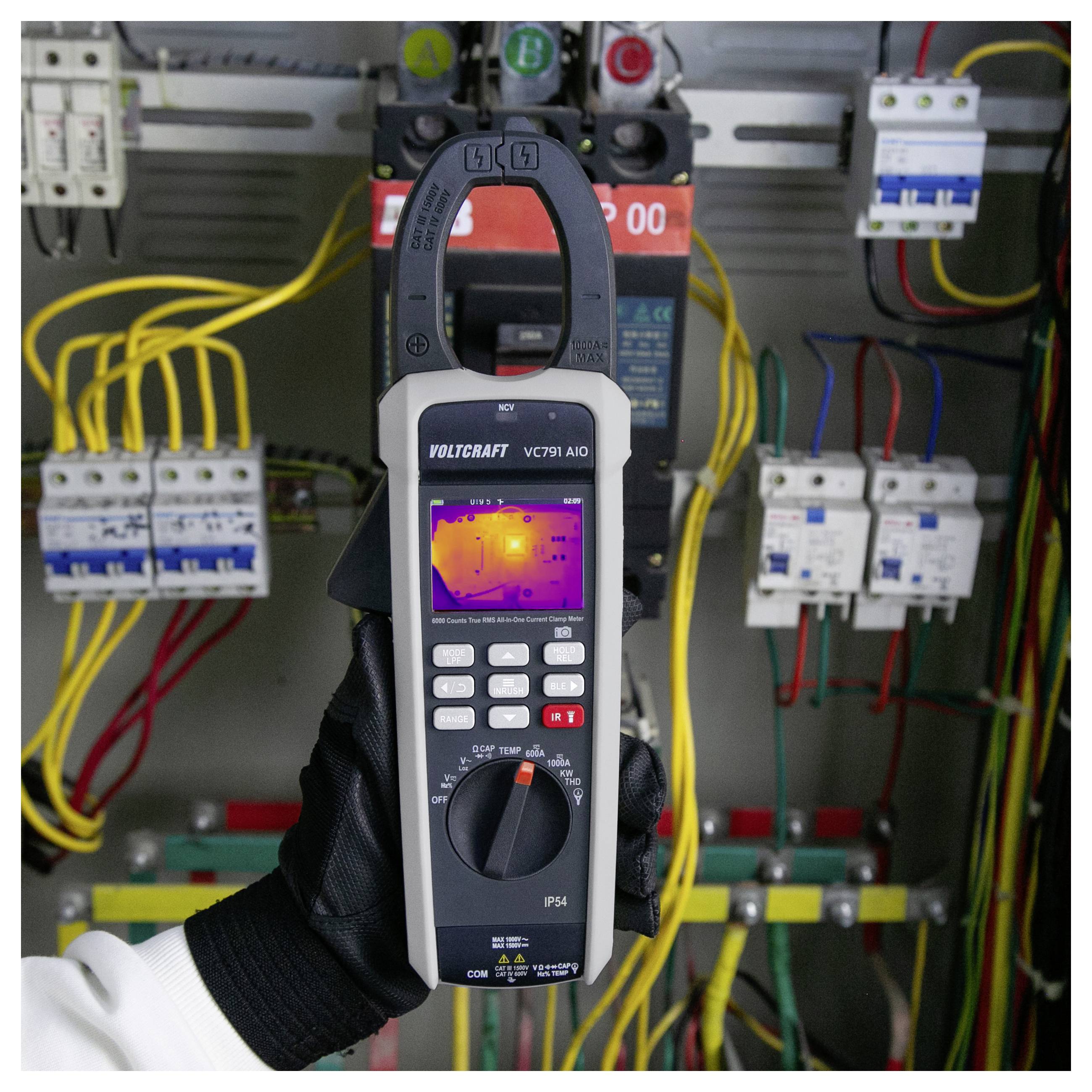 Una mano guantata tiene un multimetro a pinza VOLTCRAFT con schermo termico all'interno di un quadro elettrico di controllo con fili e interruttori automatici.