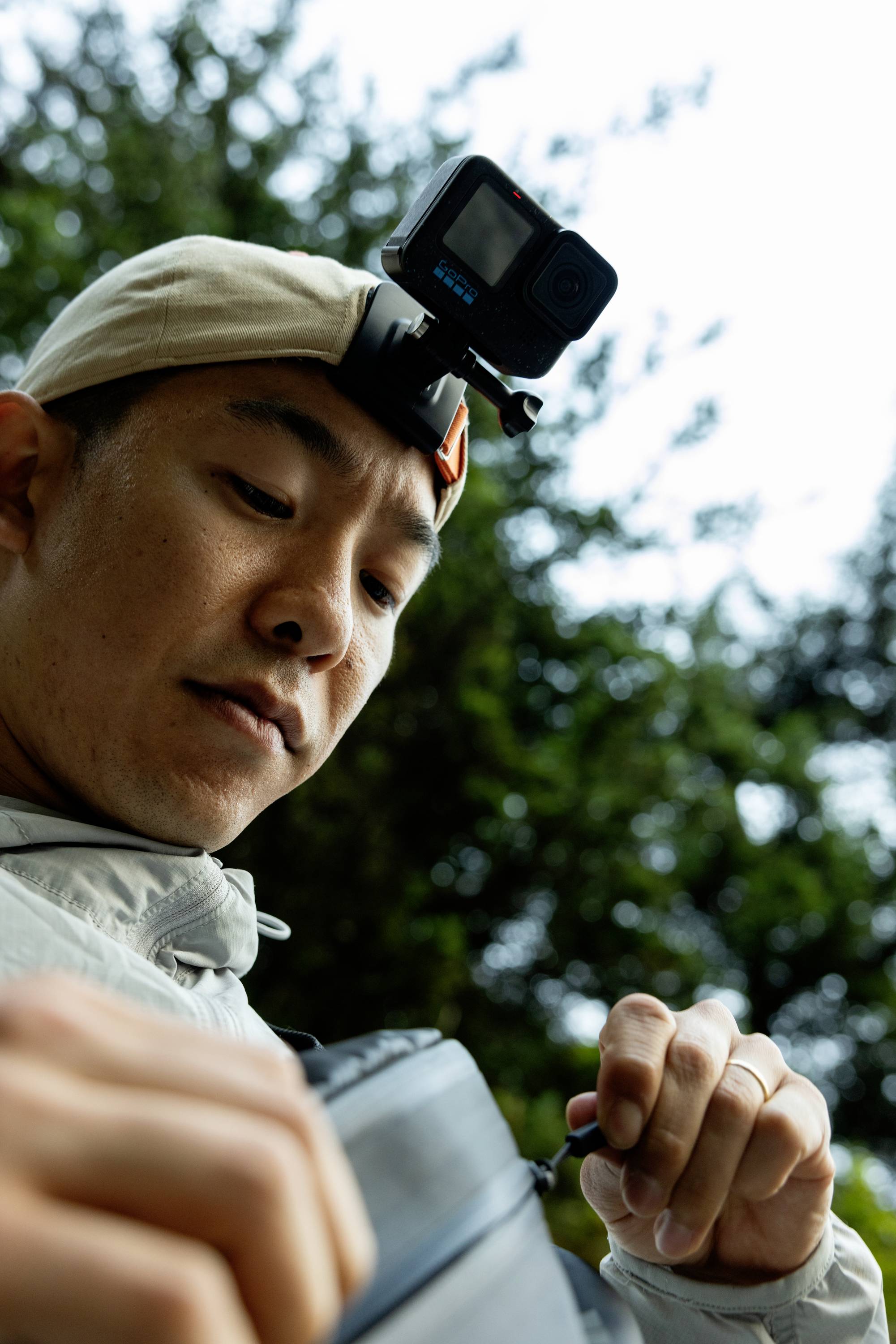 Un uomo con un cappellino bianco e una action camera sulla testa apre uno zaino, con degli alberi sullo sfondo.