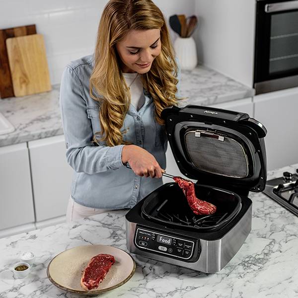 Una donna è in piedi in una cucina e sta mettendo una bistecca in una griglia elettrica. Accanto a lei sul piano di lavoro è posato un altro pezzo di carne.