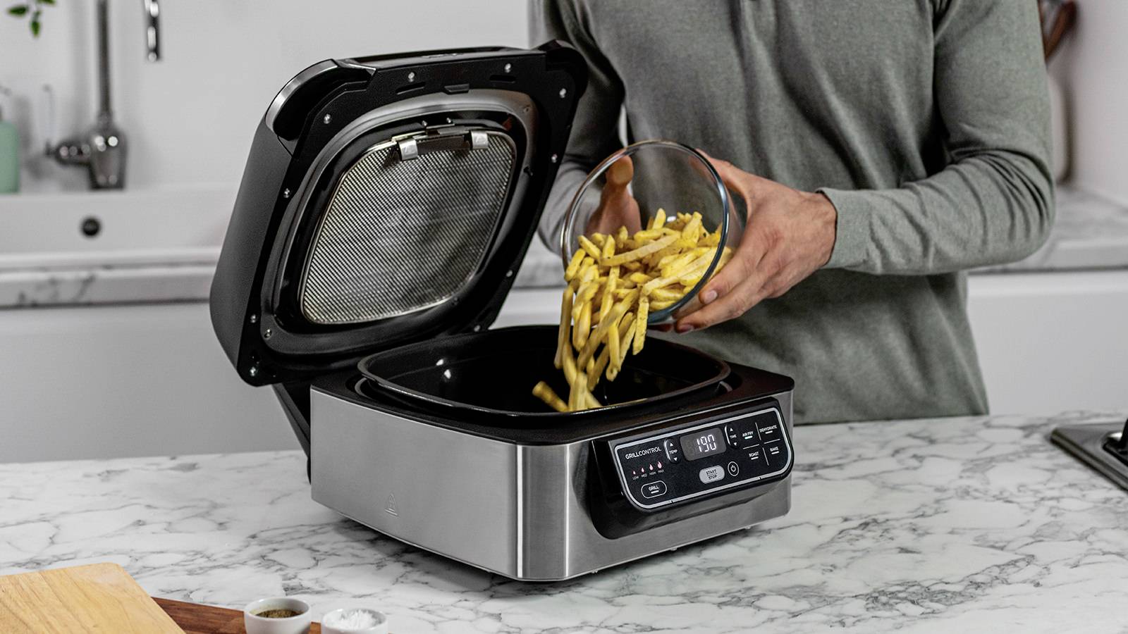 Una persona versa patatine fritte in un robot da cucina su una superficie di lavoro in marmo. La macchina ha un display digitale e il coperchio aperto.