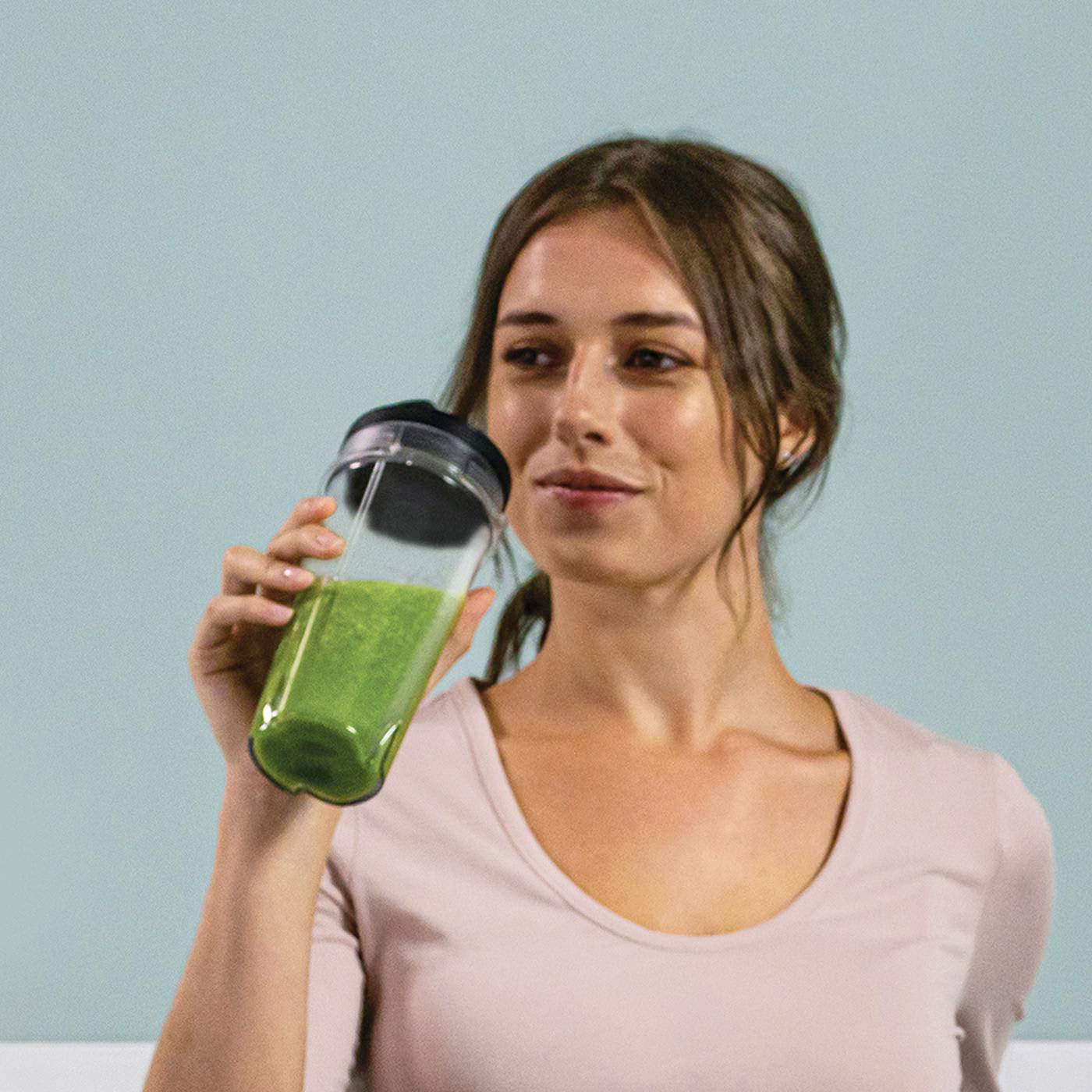 Una donna tiene in mano una tazza con uno smoothie verde e sorride. Indossa una maglia rosa e ha capelli lunghi. Lo sfondo è neutro.