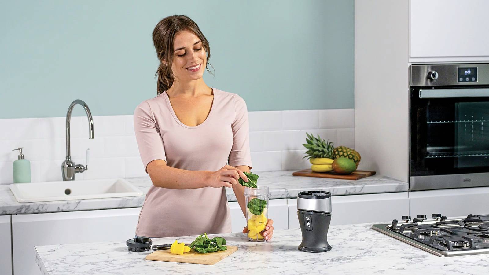 Una donna prepara un frullato in una cucina moderna. Aggiunge spinaci e frutta in un frullatore su un piano di lavoro in marmo.