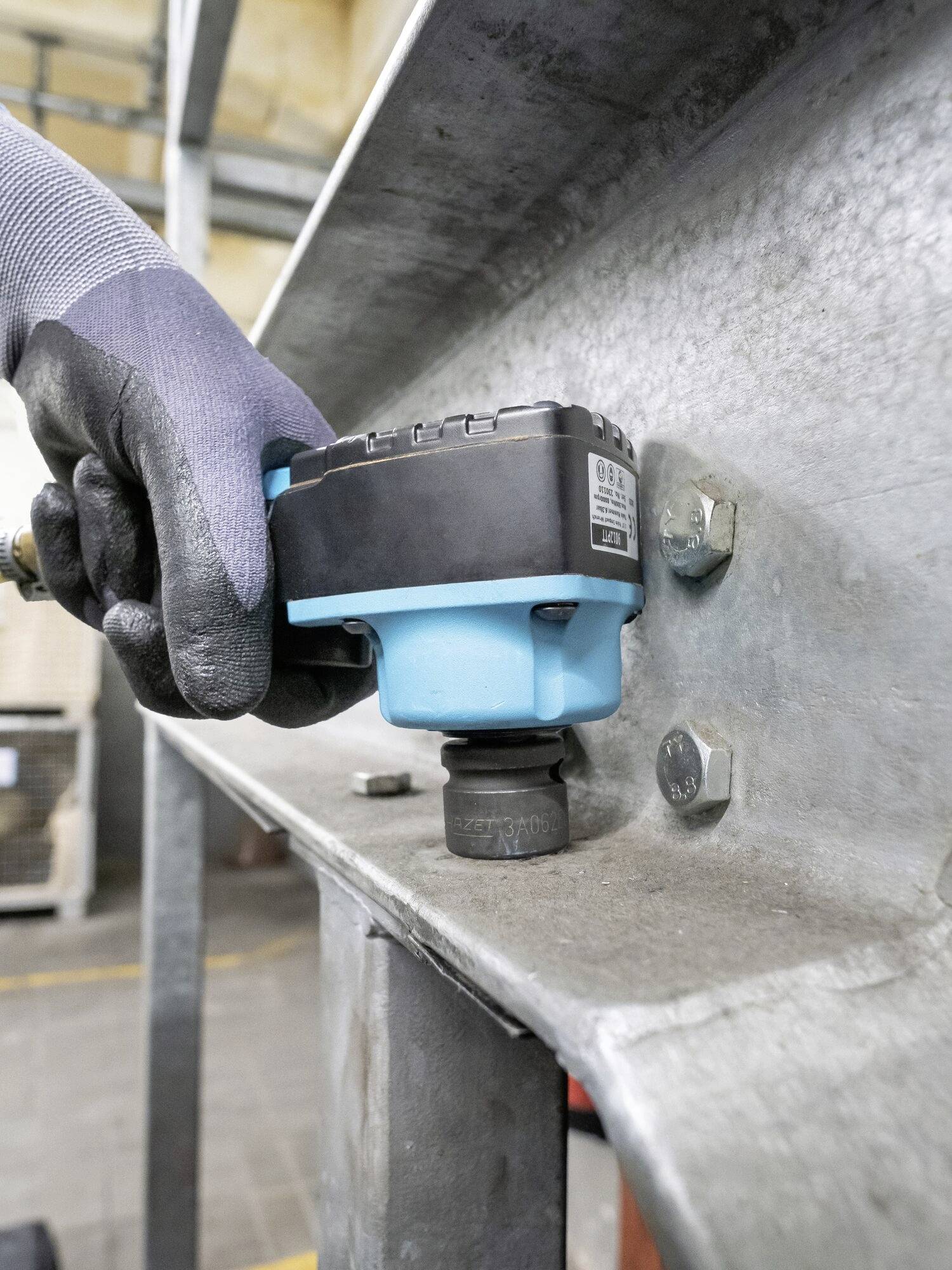 Una mano con guanti da lavoro tiene uno strumento blu e nero su un supporto metallico, che probabilmente serve per misurare o fissare qualcosa.