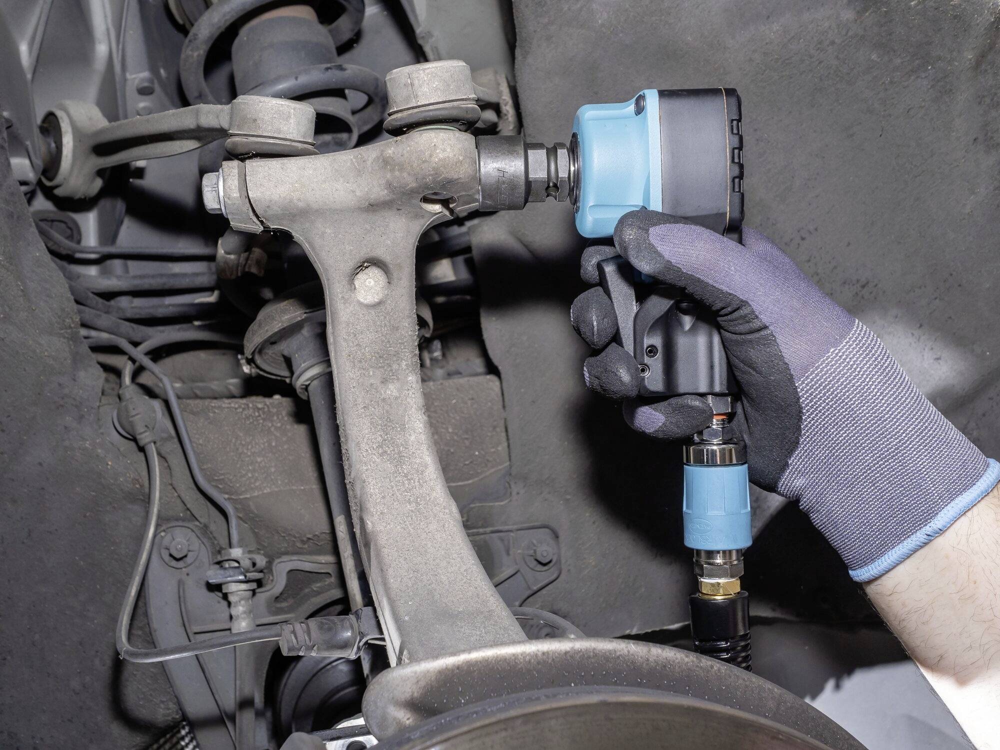 Una mano guantata tiene un utensile elettrico e serra una vite su un telaio di un veicolo, circondato da componenti automobilistici.