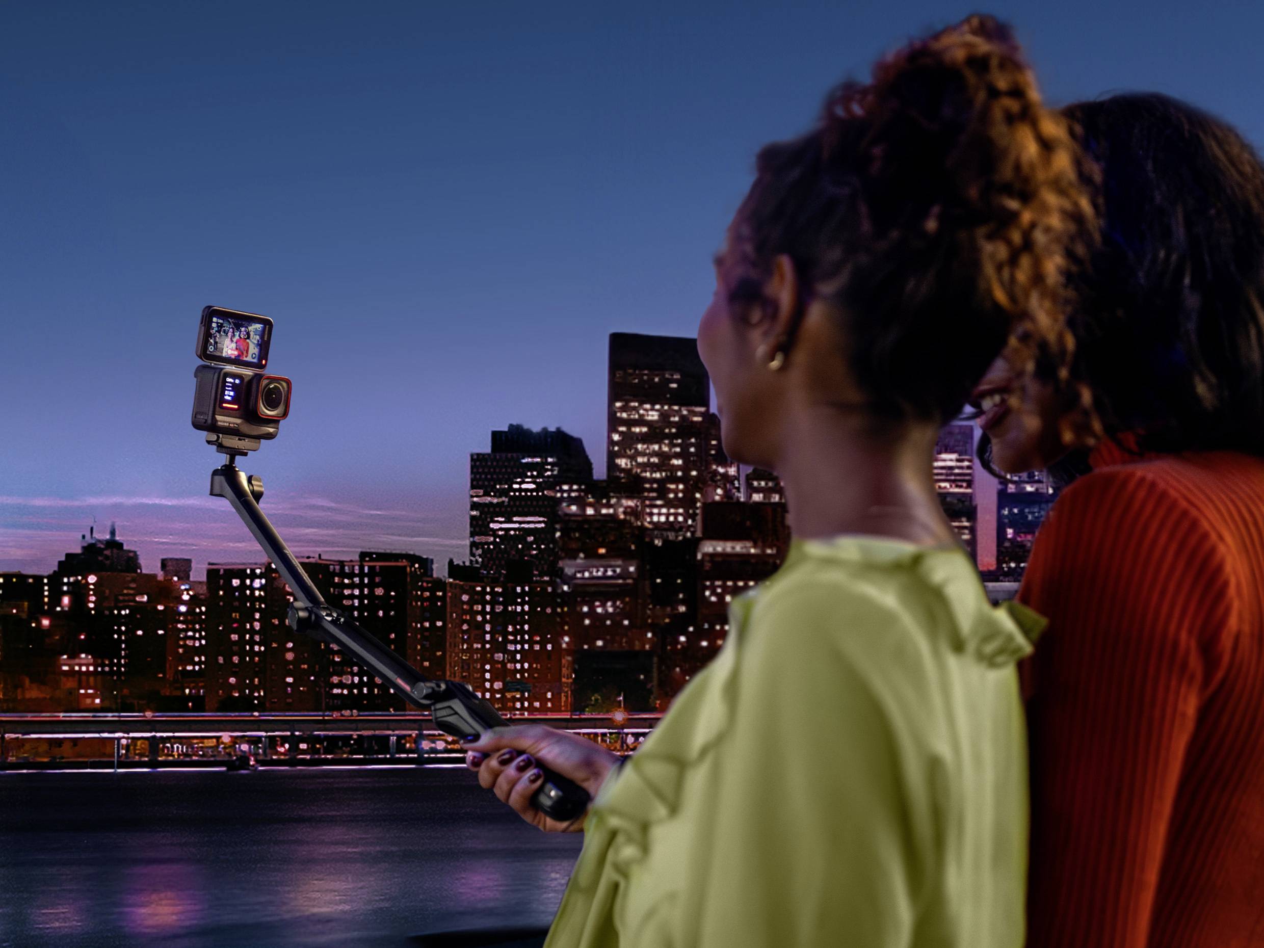 Due persone si trovano di fronte a una silhouette urbana in una serata, scattando un selfie con un selfie stick. Luci della città sullo sfondo.