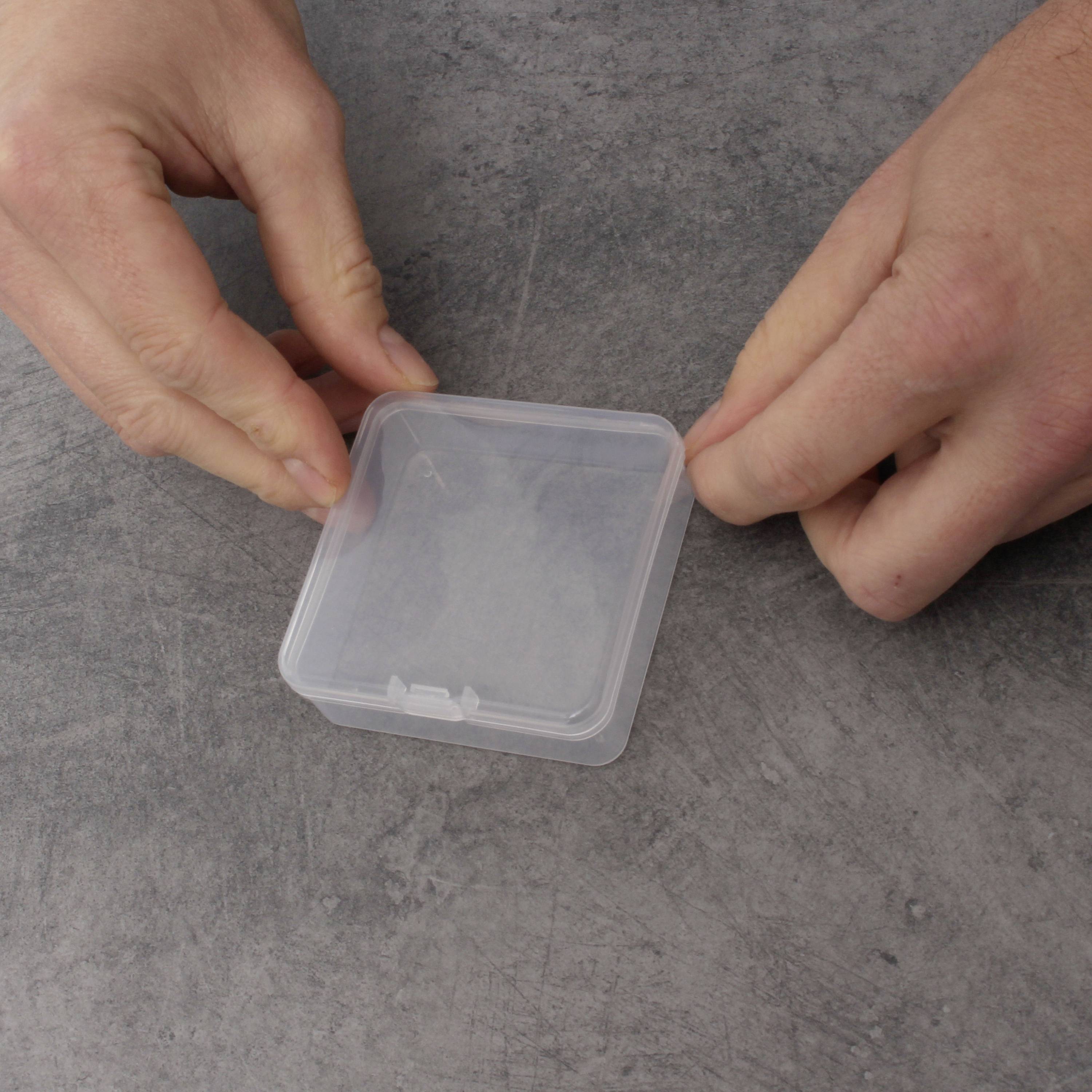 Due mani tengono una piccola scatola quadrata di plastica trasparente su una superficie di tavolo grigia.
