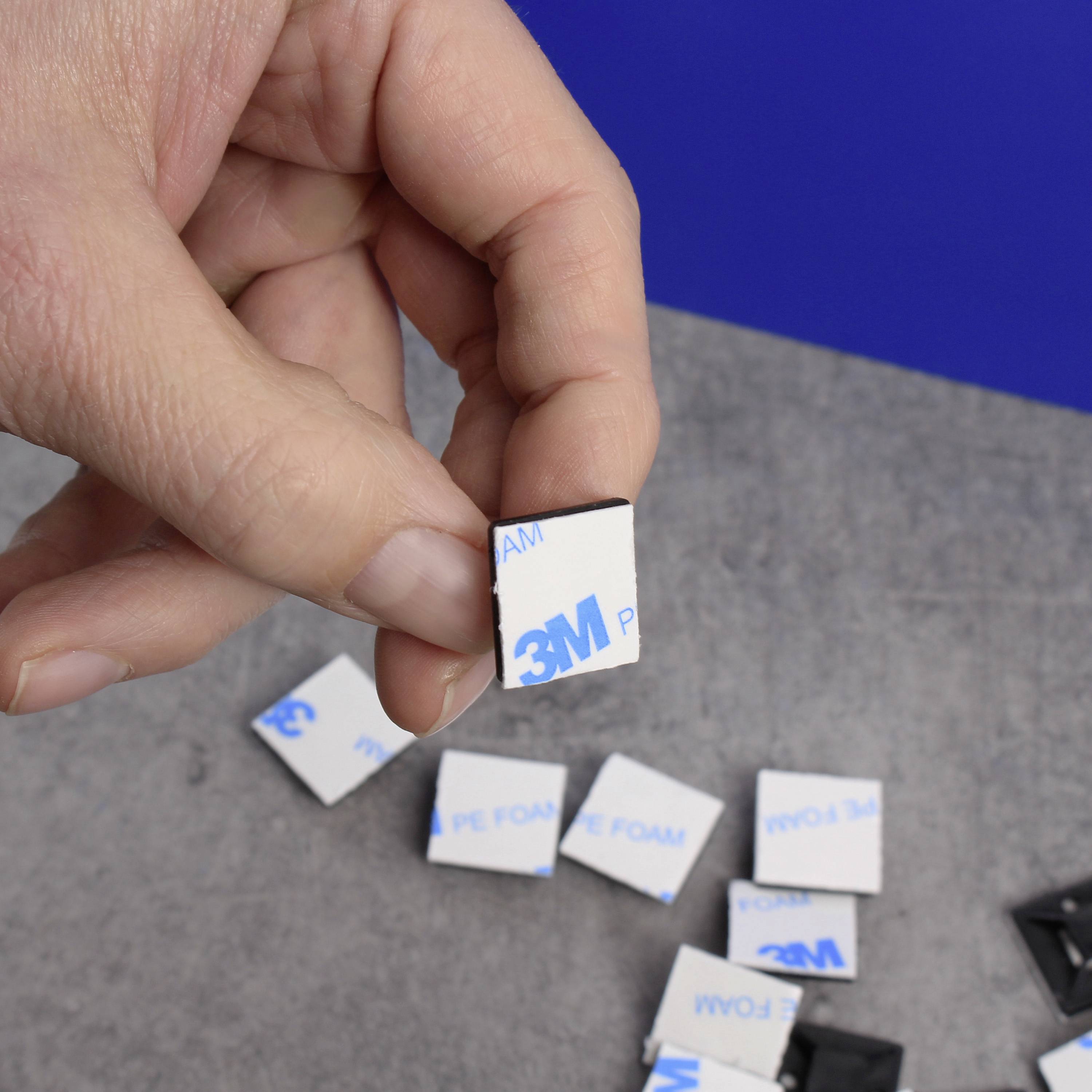 Una mano tiene un pad adesivo quadrato blu e bianco con il logo '3M'. Diversi pad simili sono posizionati su un tavolo grigio.
