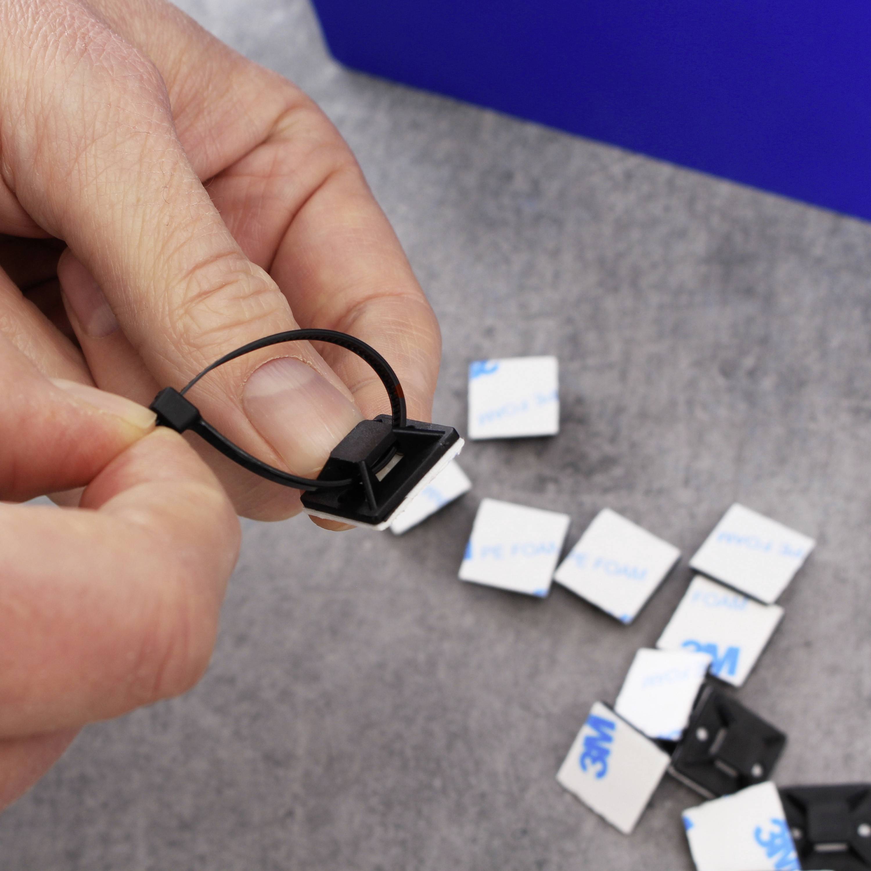 Una mano tira una fascetta per cavi nera attraverso un supporto, con piccoli pezzi quadrati di nastro adesivo sullo sfondo.