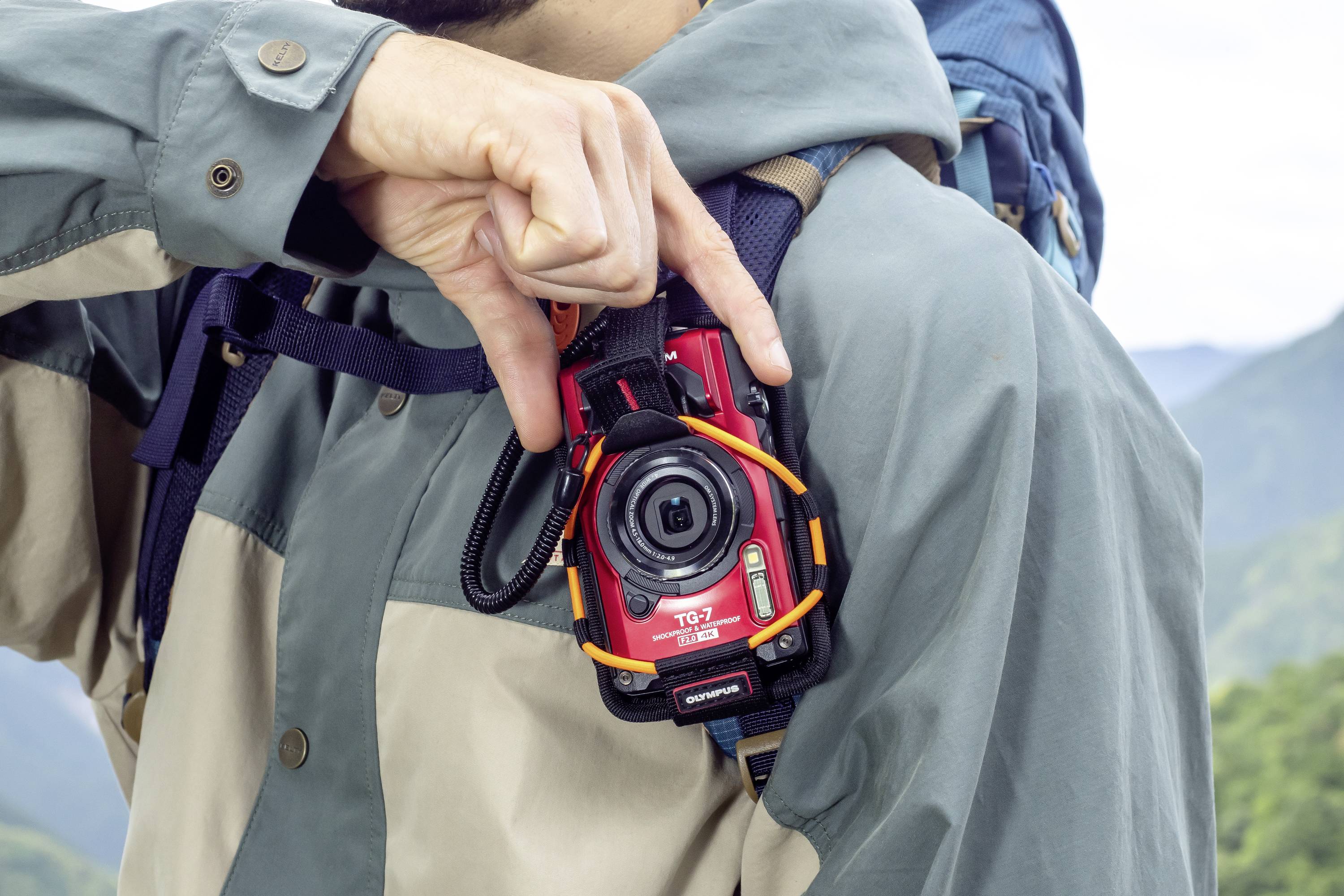 Un uomo vestito con abbigliamento da esterno porta una fotocamera rossa al collo e fa un gesto con la mano. Sullo sfondo sono visibili delle montagne.
