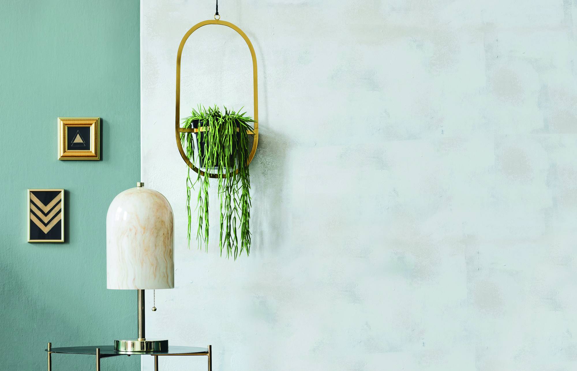 Stanza minimalista con una lampada bianca, una pianta sospesa e due quadri incorniciati appesi su una parete color turchese e bianco pallido.