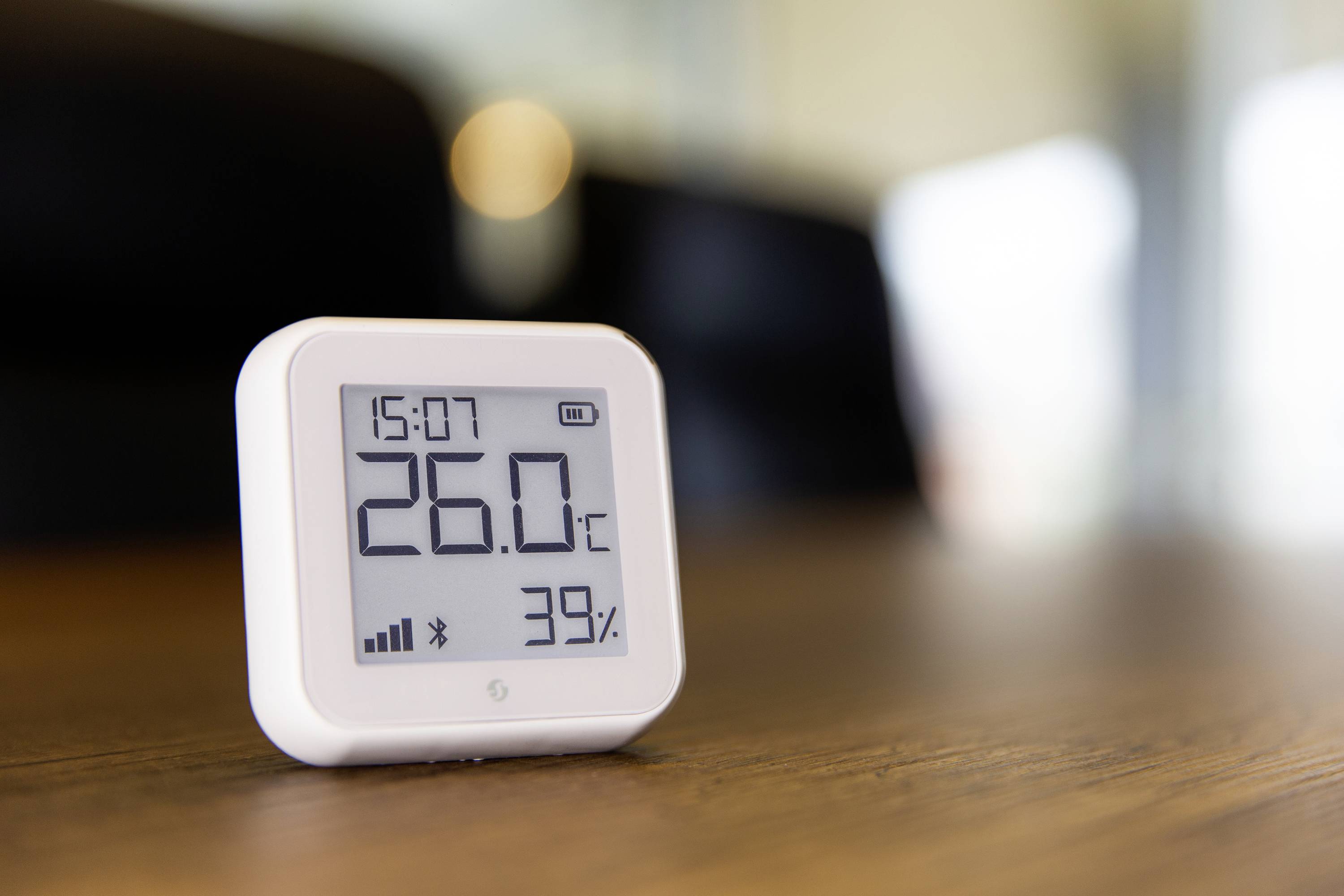 Un termometro digitale mostra 26,0 °C e il 39% di umidità su un tavolo di legno, con una stanza sullo sfondo.