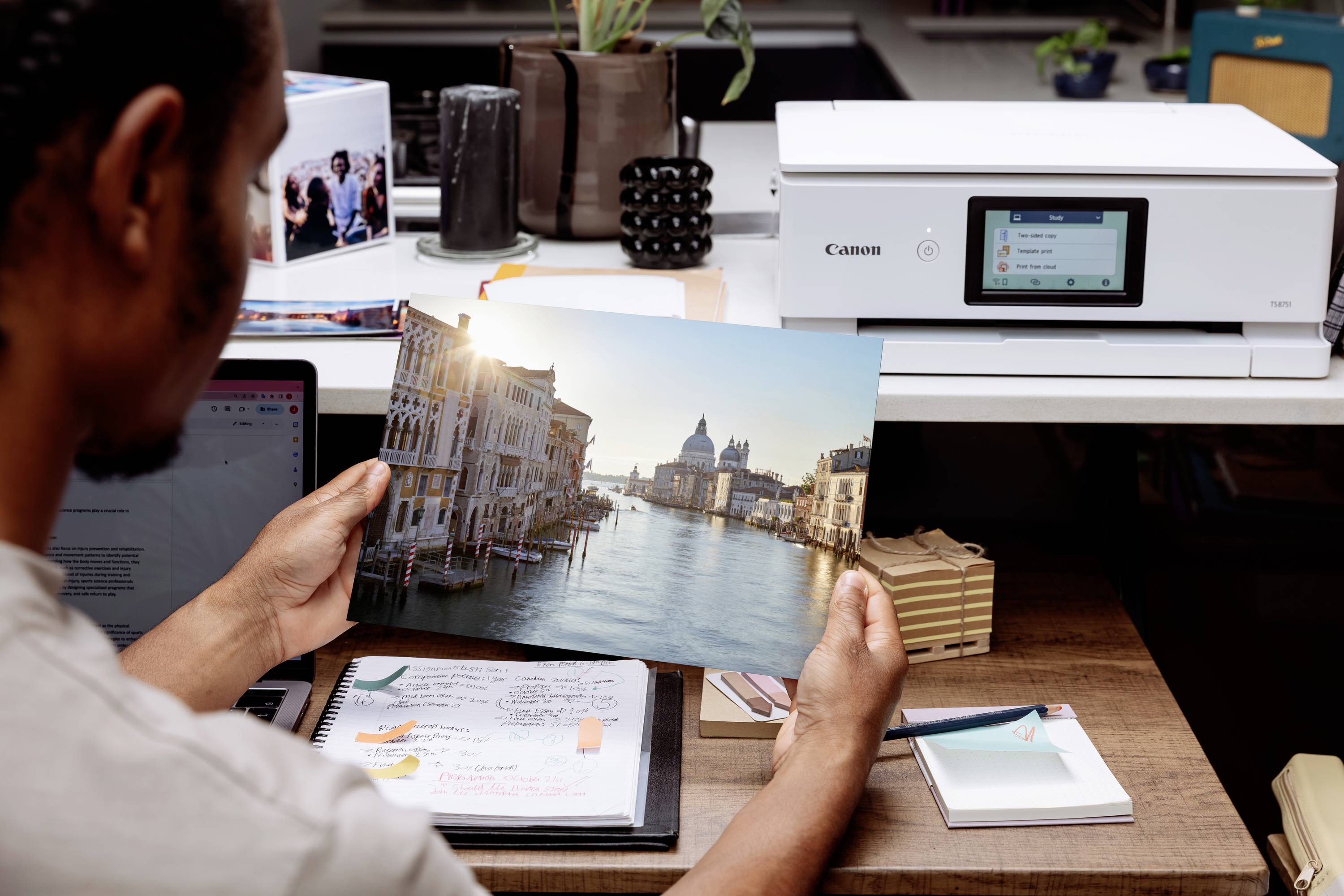 Una persona sta osservando una foto di Venezia, appena stampata con una stampante su una scrivania. Sullo sfondo ci sono piante e materiale da ufficio.