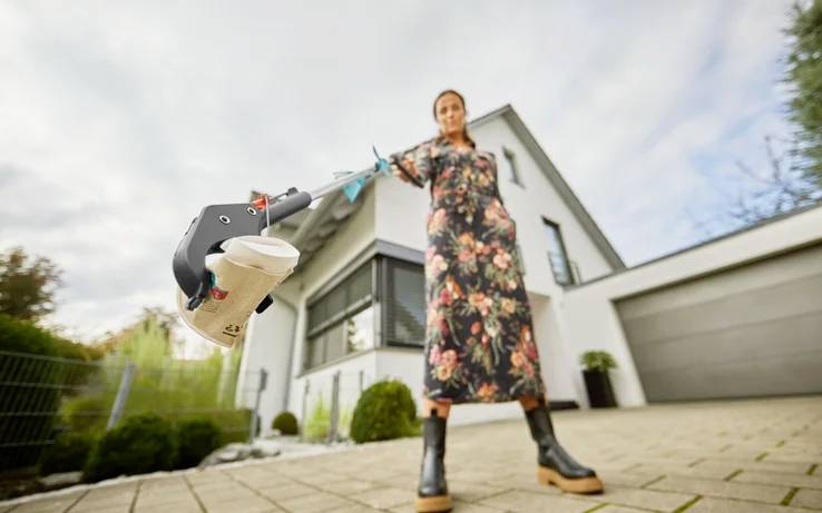 Donna in un abito lungo con stampa floreale è in piedi davanti a una casa moderna e raccoglie la spazzatura con un pinzatore rivolto in avanti.