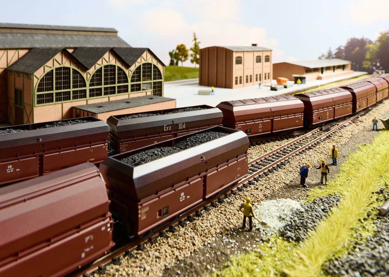 Scena ferroviaria in miniatura con diversi carri merci carichi di carbone sui binari davanti a un edificio della stazione, circondati da piccole figure umane.