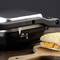 Un panino con formaggio fuso viene pressato in una piastra per toast. È adagiato su un tagliere di legno ed è parzialmente visibile.