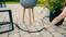 Una persona sistema un tubo da giardino nero su una terrazza in pietra, circondato da piante in vasi moderni, suggerendo attività di giardinaggio all'aperto.