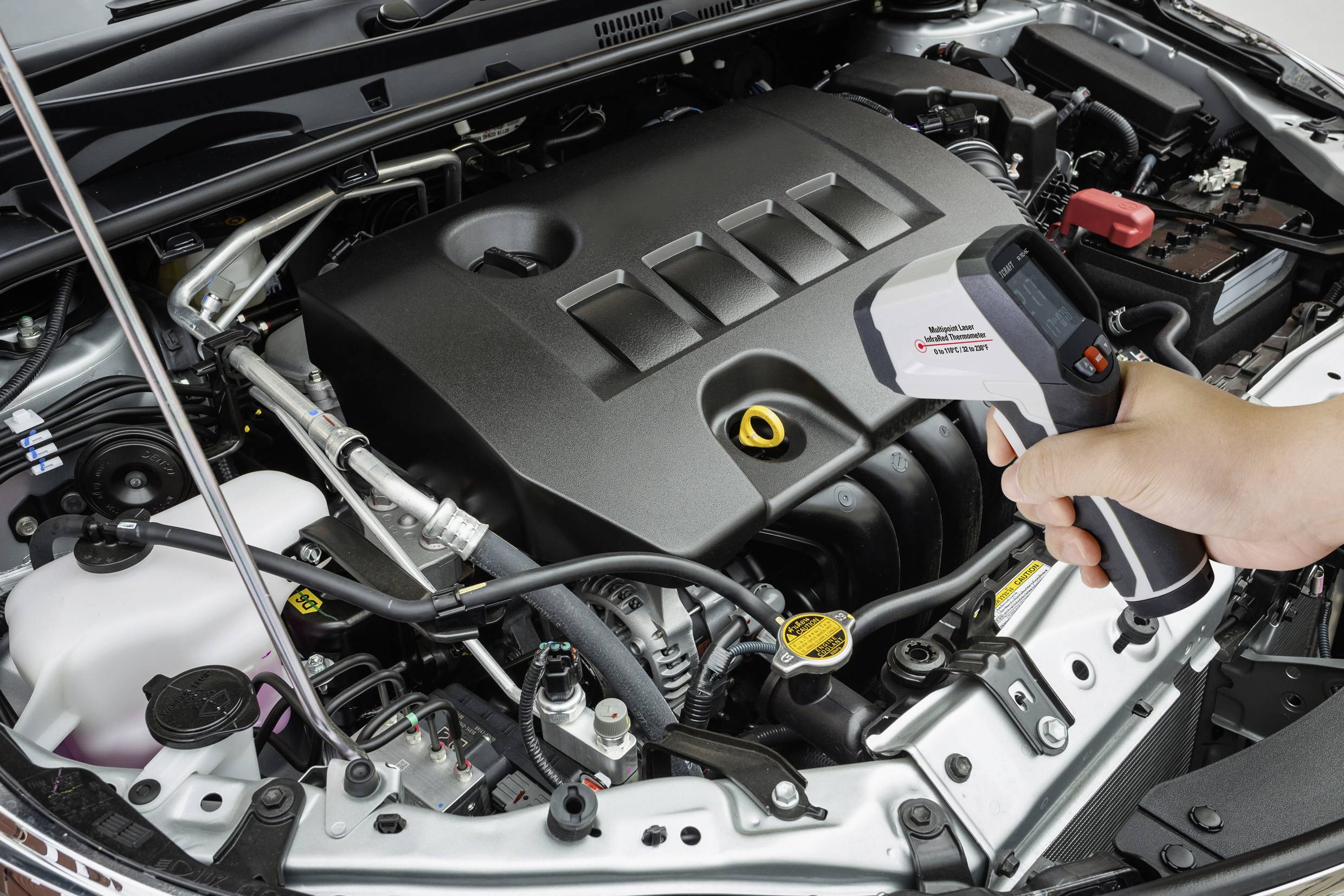 Una persona tiene un termometro digitale sopra il vano motore aperto di un'auto. Lo strumento di misurazione indica 35,2 gradi Celsius.