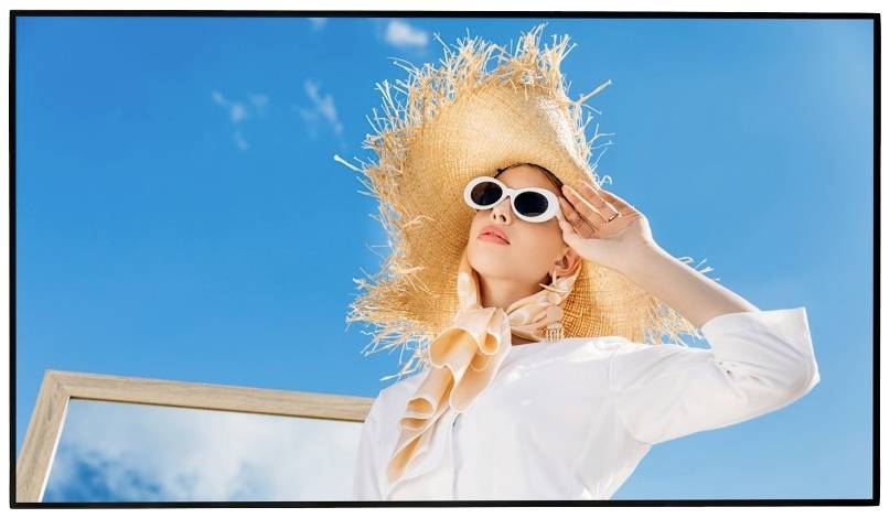 Donna con cappello di paglia e occhiali da sole posa davanti a un cielo blu, indossa una camicia bianca. Tiene fermo il cappello con la sua mano destra.