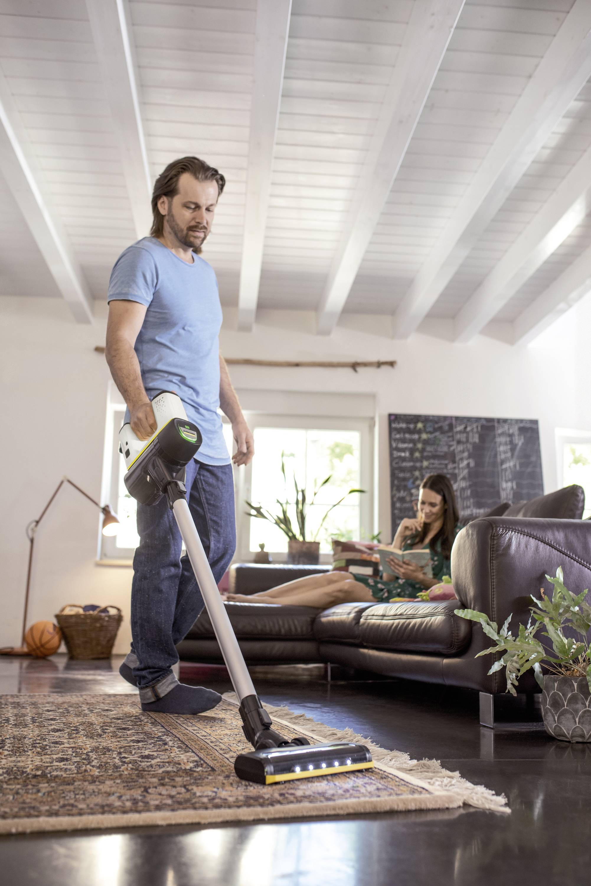 Un uomo aspira il tappeto in un soggiorno arredato in stile moderno, mentre una donna è seduta rilassata sul divano.