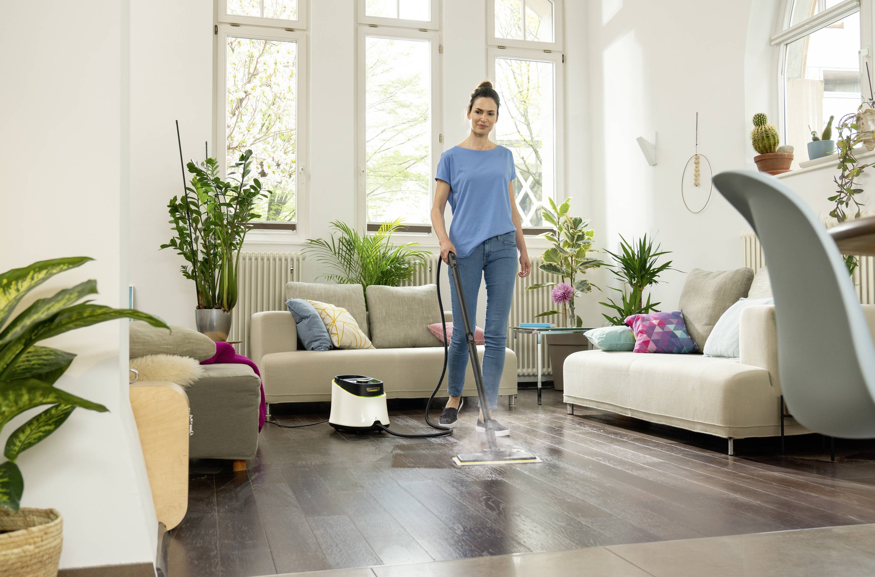 Una donna pulisce il pavimento di un soggiorno accogliente con un pulitore a vapore. L'ambiente è luminoso e decorato con piante.