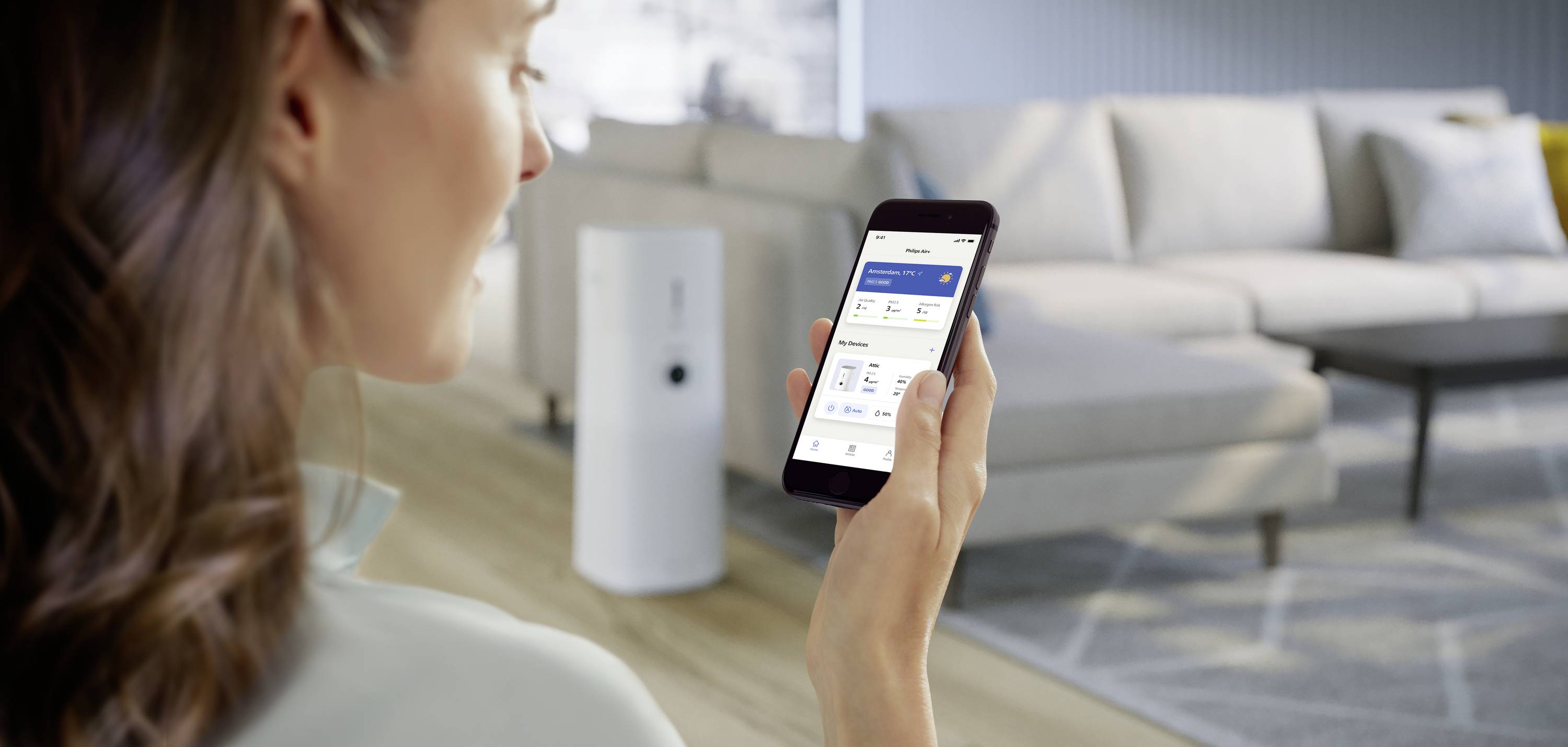 Una persona tiene in mano uno smartphone con un'app aperta, con mobili imbottiti e un purificatore d'aria sullo sfondo. Contesto: controllo domestico intelligente.