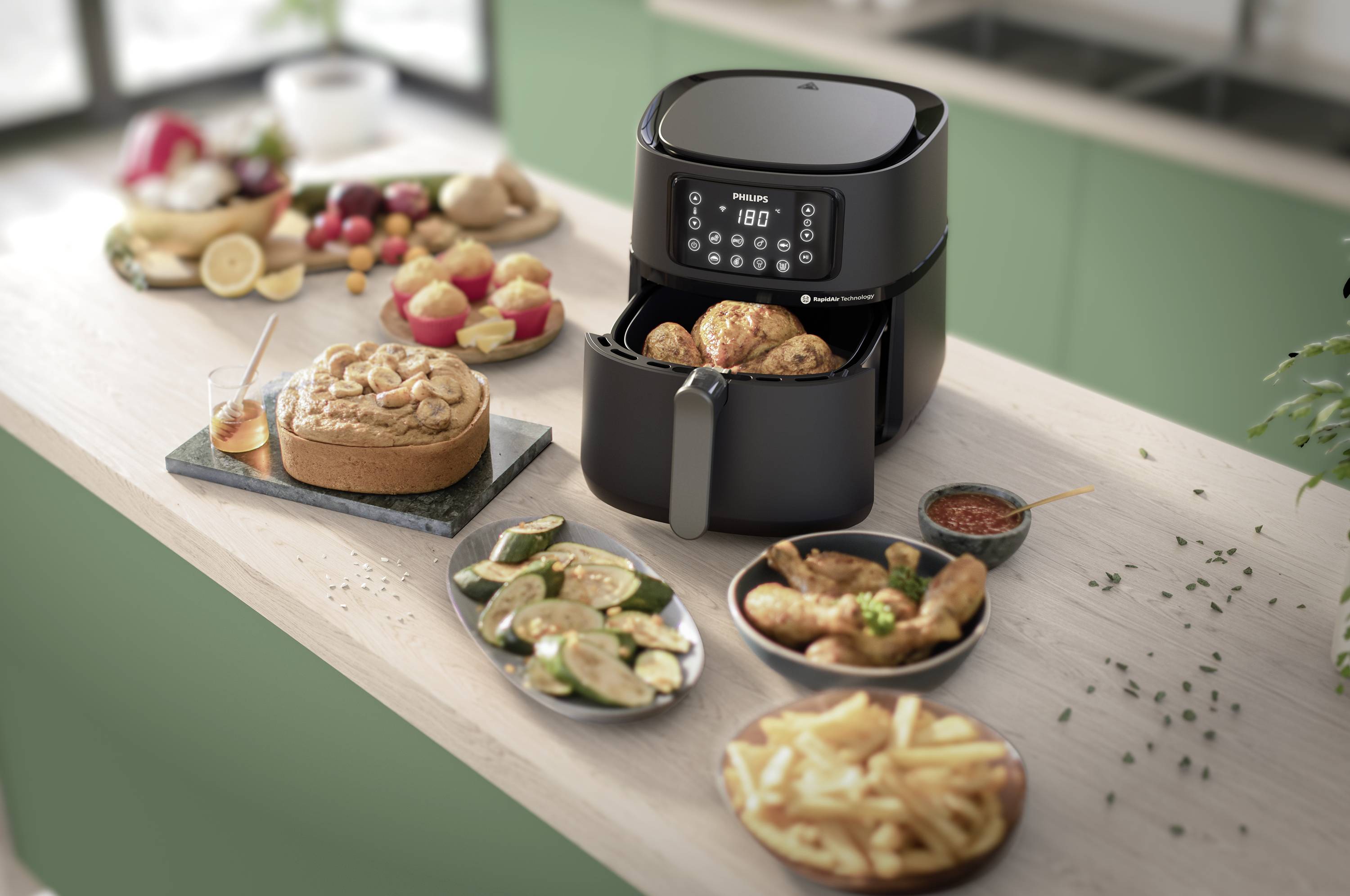 Una friggitrice ad aria calda è posizionata su un piano di lavoro della cucina, circondata da vari cibi preparati come patatine fritte, cosce di pollo e torta.