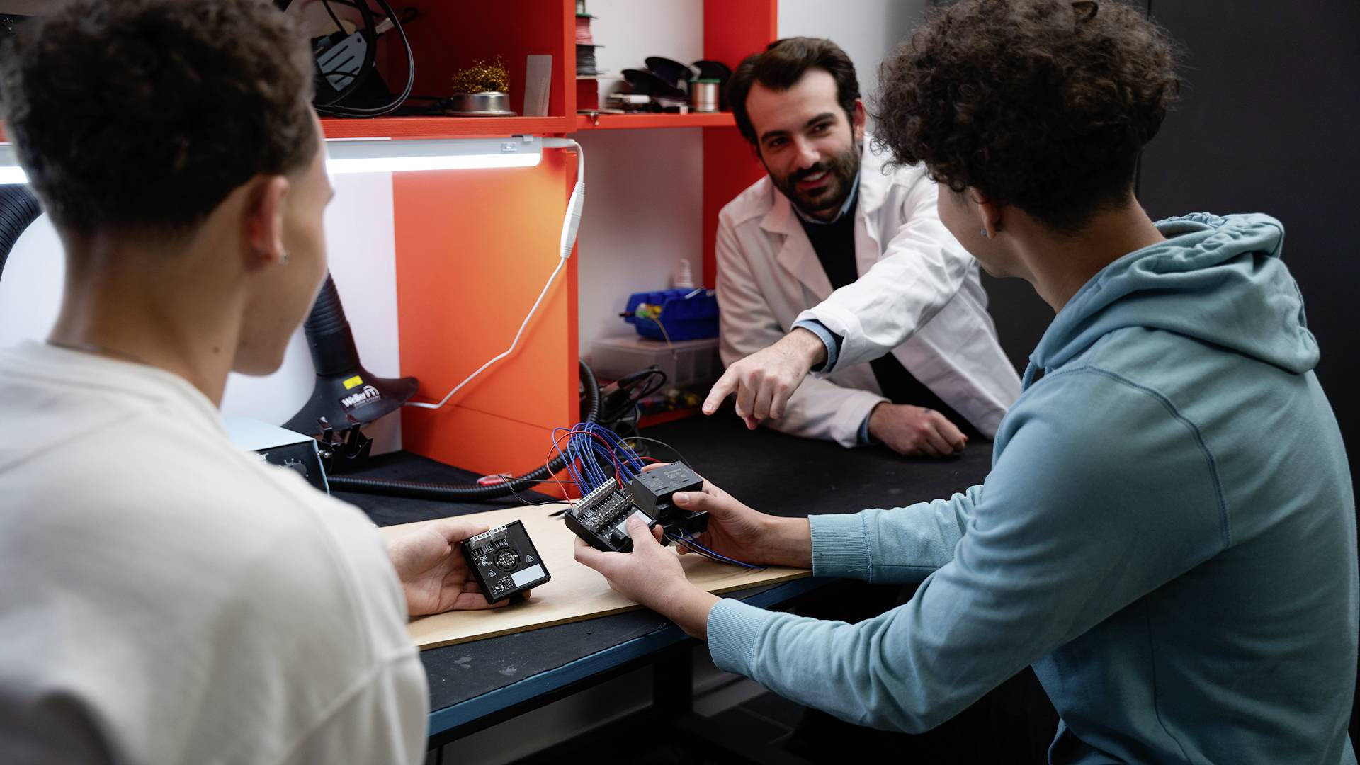 Tre persone in un laboratorio. Due giovani tengono dispositivi elettronici mentre un uomo in camice bianco spiega.