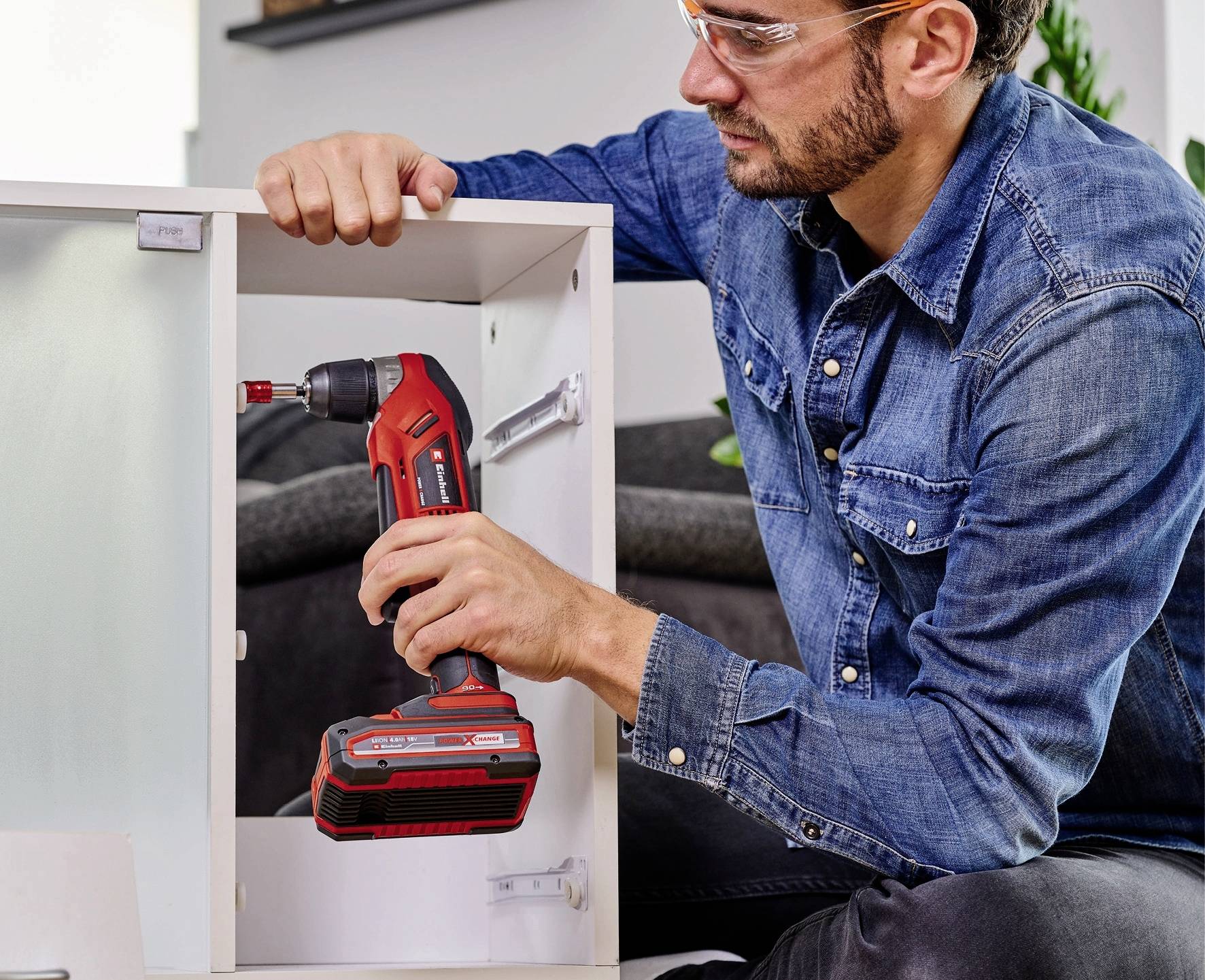 Un uomo con una camicia di jeans sta usando un cacciavite a batteria rosso per assemblare un mobile, lavorando in modo concentrato e accurato.