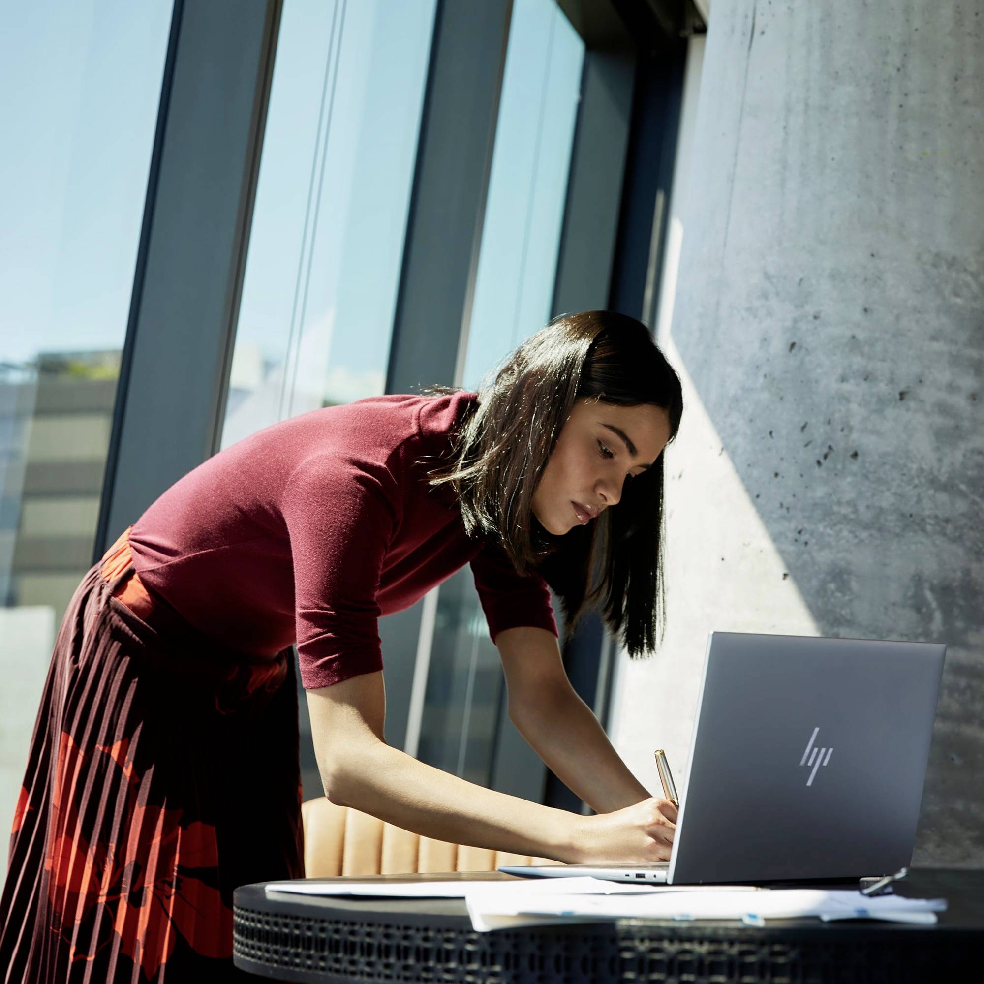 Una donna scrive in un quaderno mentre è seduta a un tavolo con un laptop. L'ambiente mostra un ufficio moderno con finestre.