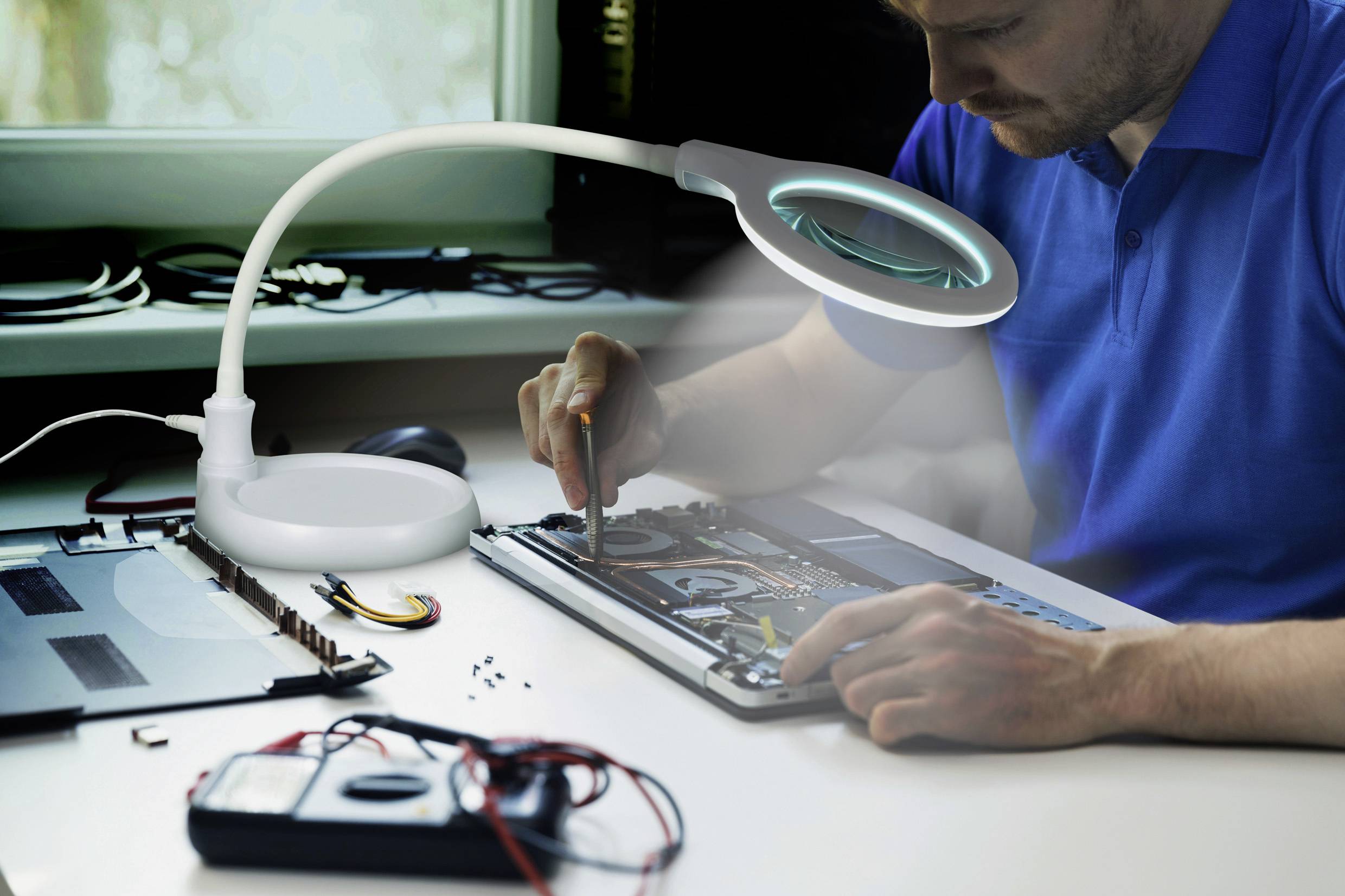 Un uomo ripara un laptop a un tavolo sotto una lente d'ingrandimento. Nel primo piano sono posizionati degli strumenti elettronici.