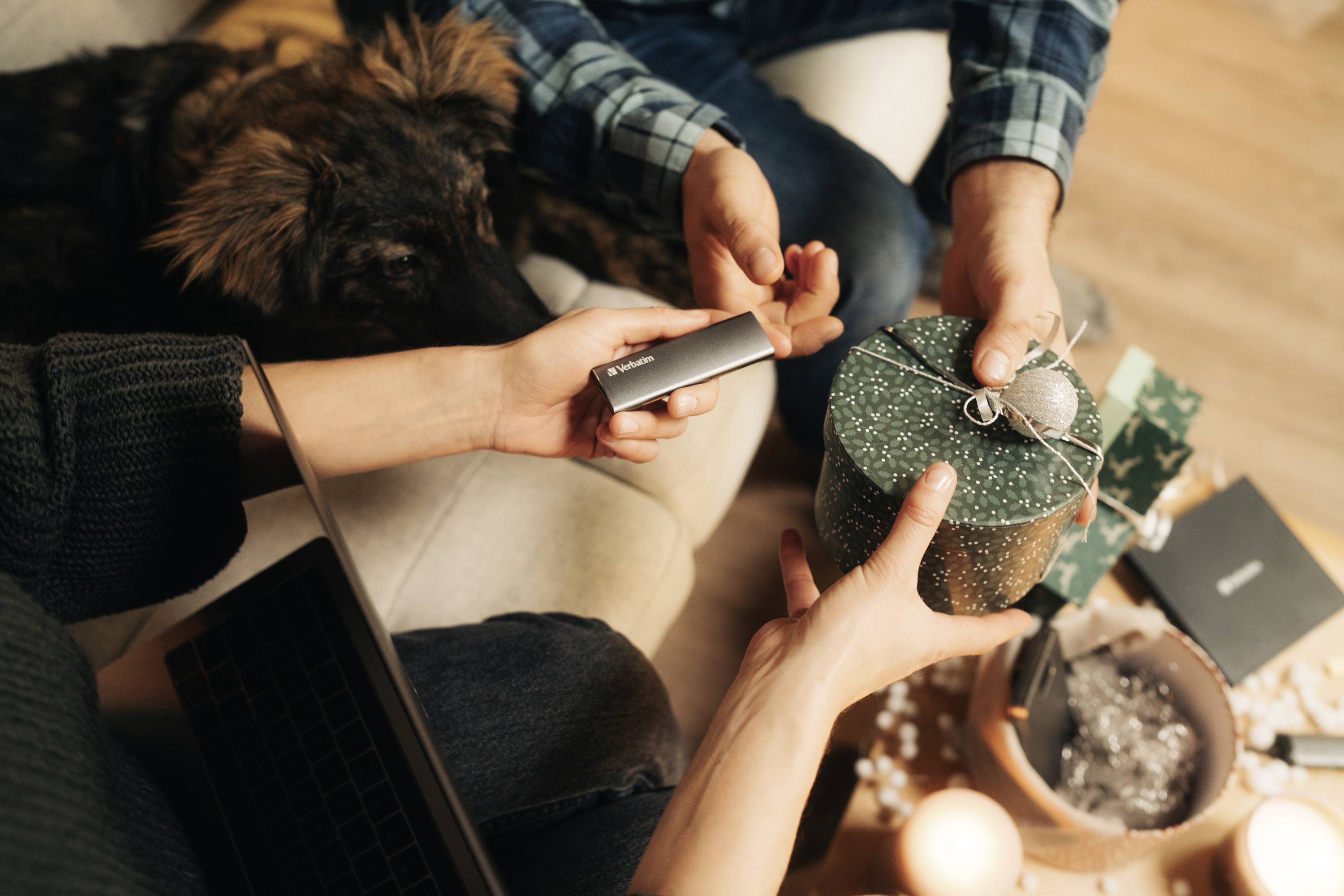 Persone si scambiano regali mentre un cane osserva. Una persona tiene in mano una scatola rotonda, un'altra sta consegnando una chiavetta USB.