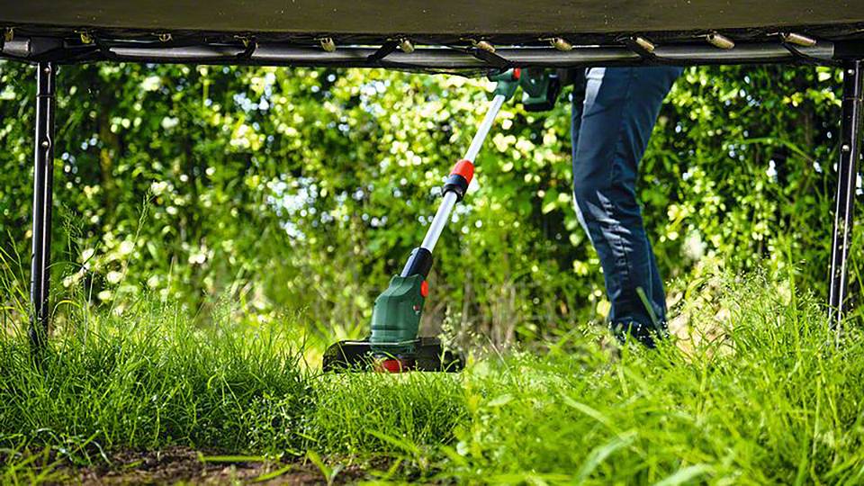 Uomo taglia l'erba sotto un trampolino con un decespugliatore elettrico; cespugli verdi sullo sfondo.
