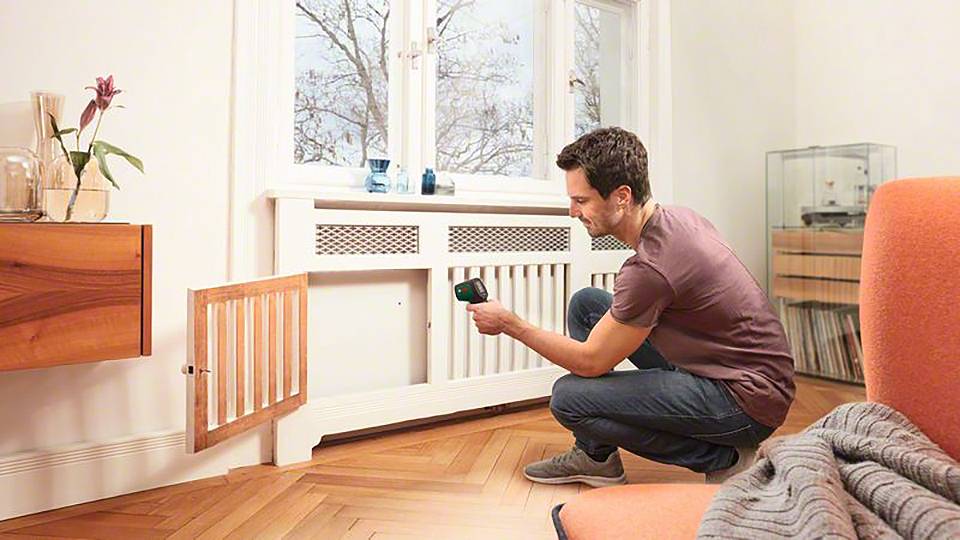 Una persona controlla la temperatura di un termosifone con un termometro a infrarossi in un soggiorno con pavimento in legno e una finestra sullo sfondo.