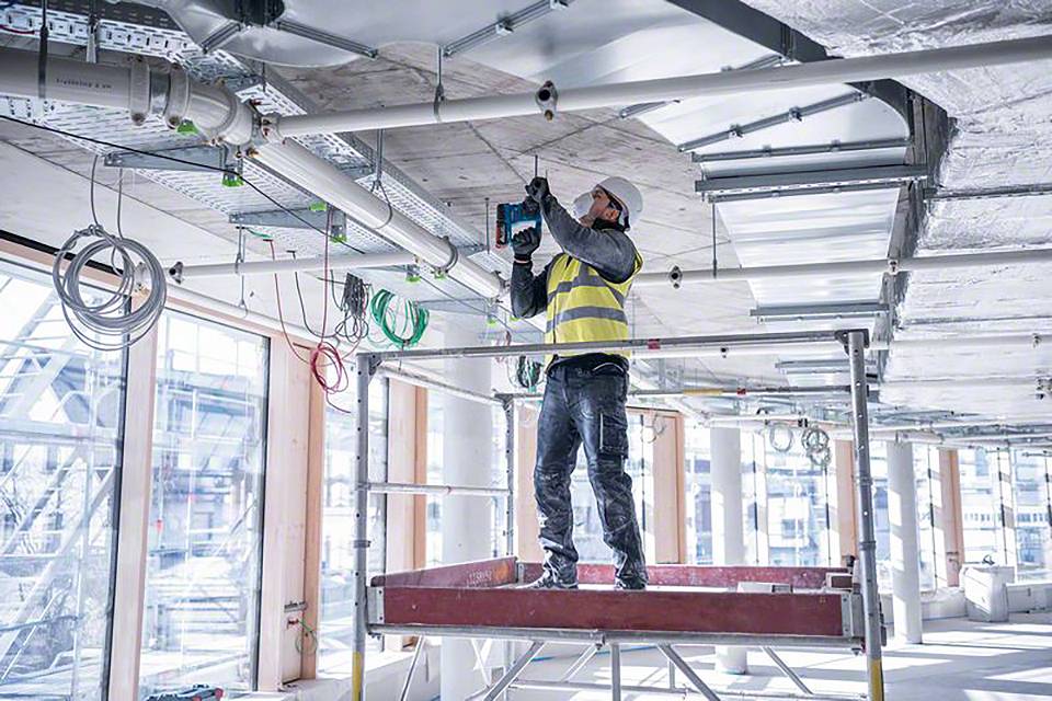 Un lavoratore edile con indumenti protettivi è in piedi su un ponteggio e sta eseguendo lavori di installazione sul soffitto di un edificio non ancora completato.
