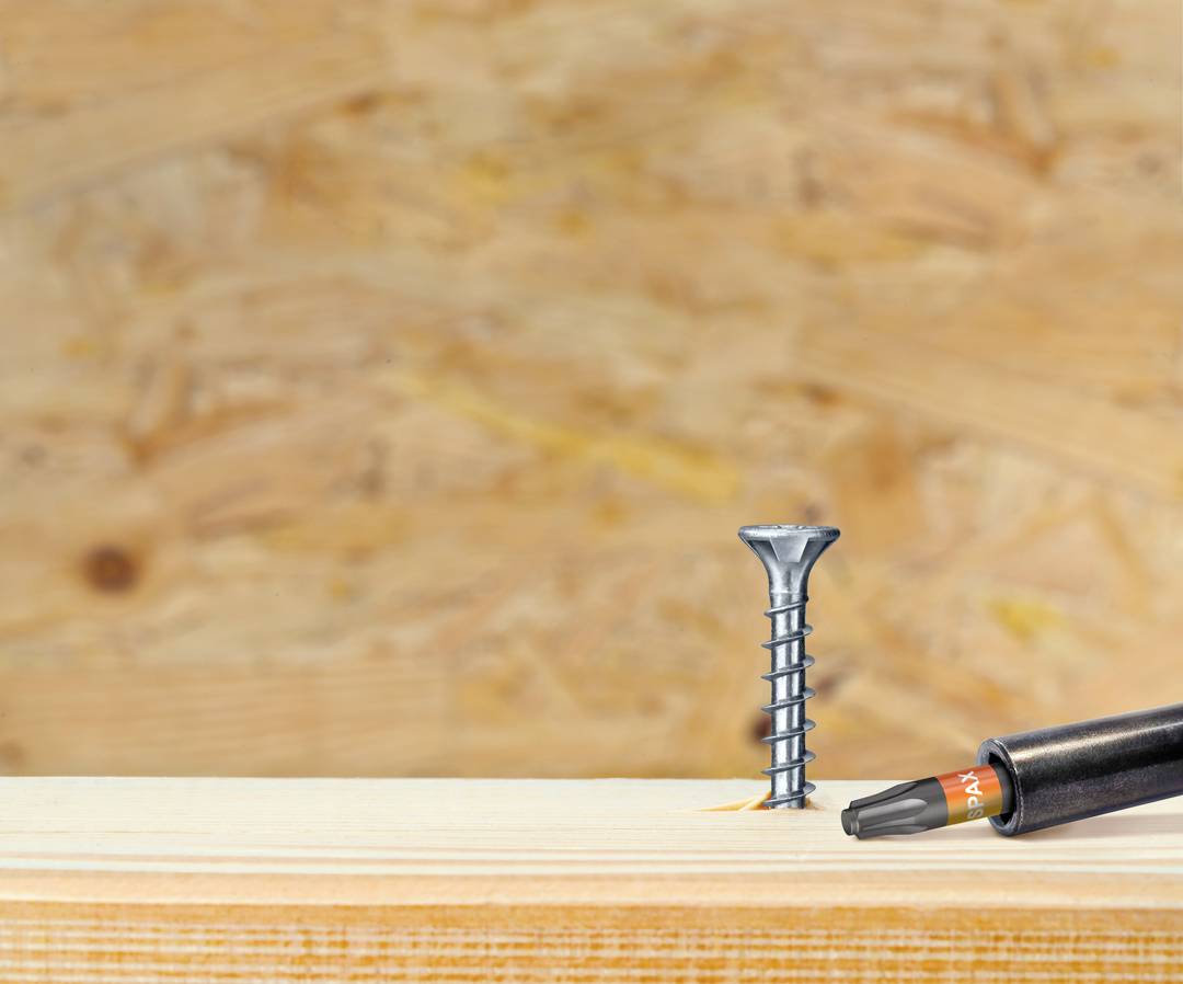 Una vite è dritta accanto a un cacciavite su una tavola di legno, davanti a uno sfondo sfocato con venature del legno.