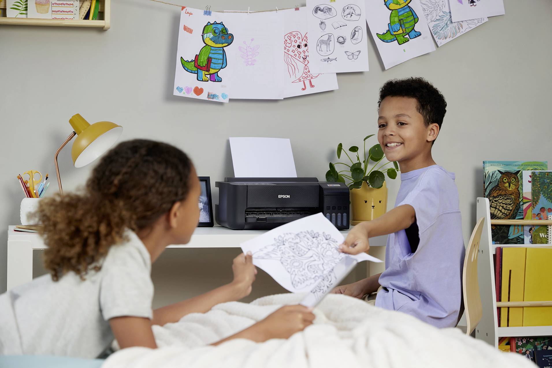 Bambini disegnano immagini sorridendo e utilizzando una stampante in un ambiente di lavoro luminoso e creativo. I disegni sono appesi alle pareti.