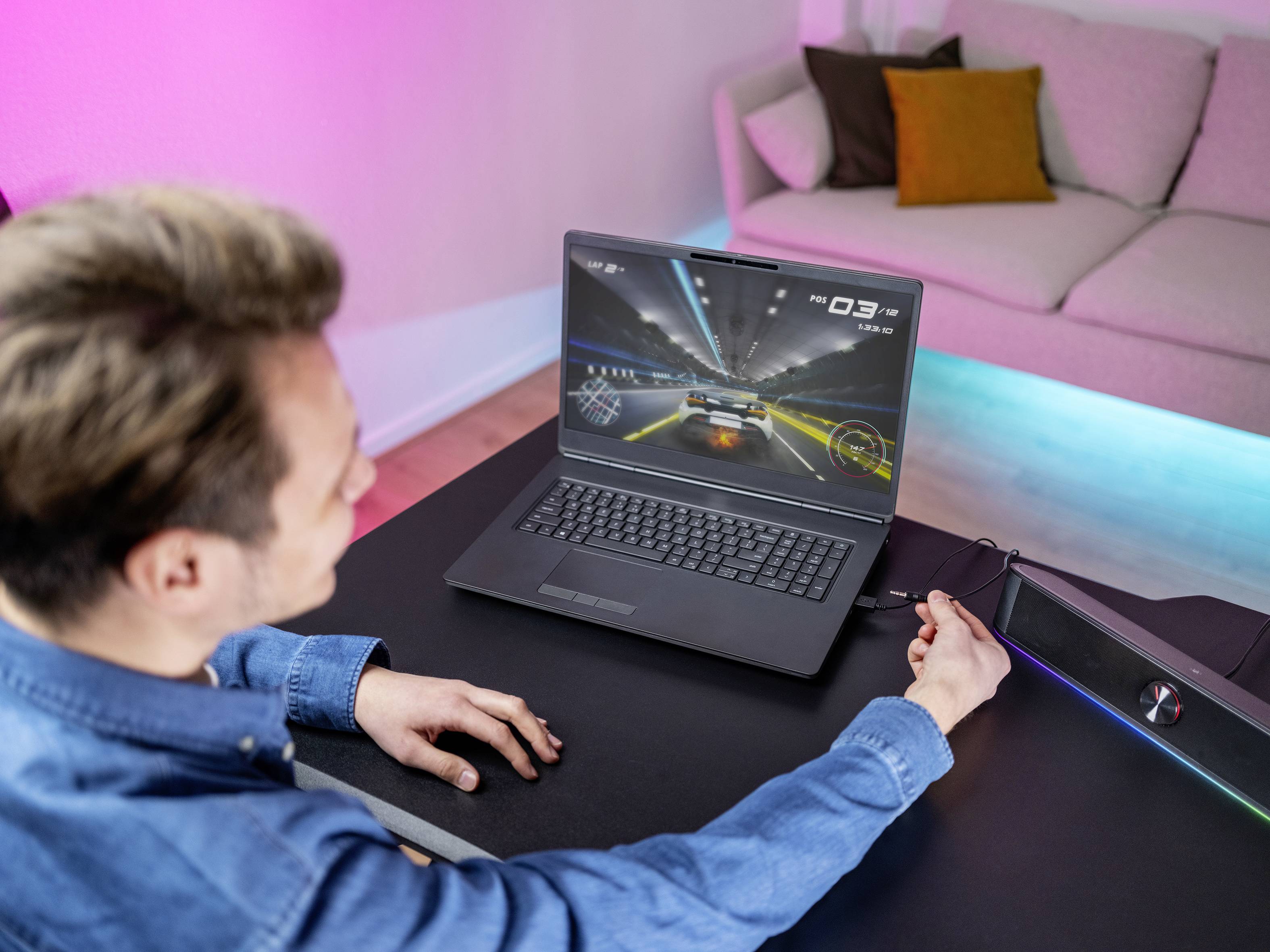 Un uomo sta giocando a un videogioco di corse automobilistiche su un laptop. Sullo sfondo si vede un divano. La stanza è illuminata da luci colorate.