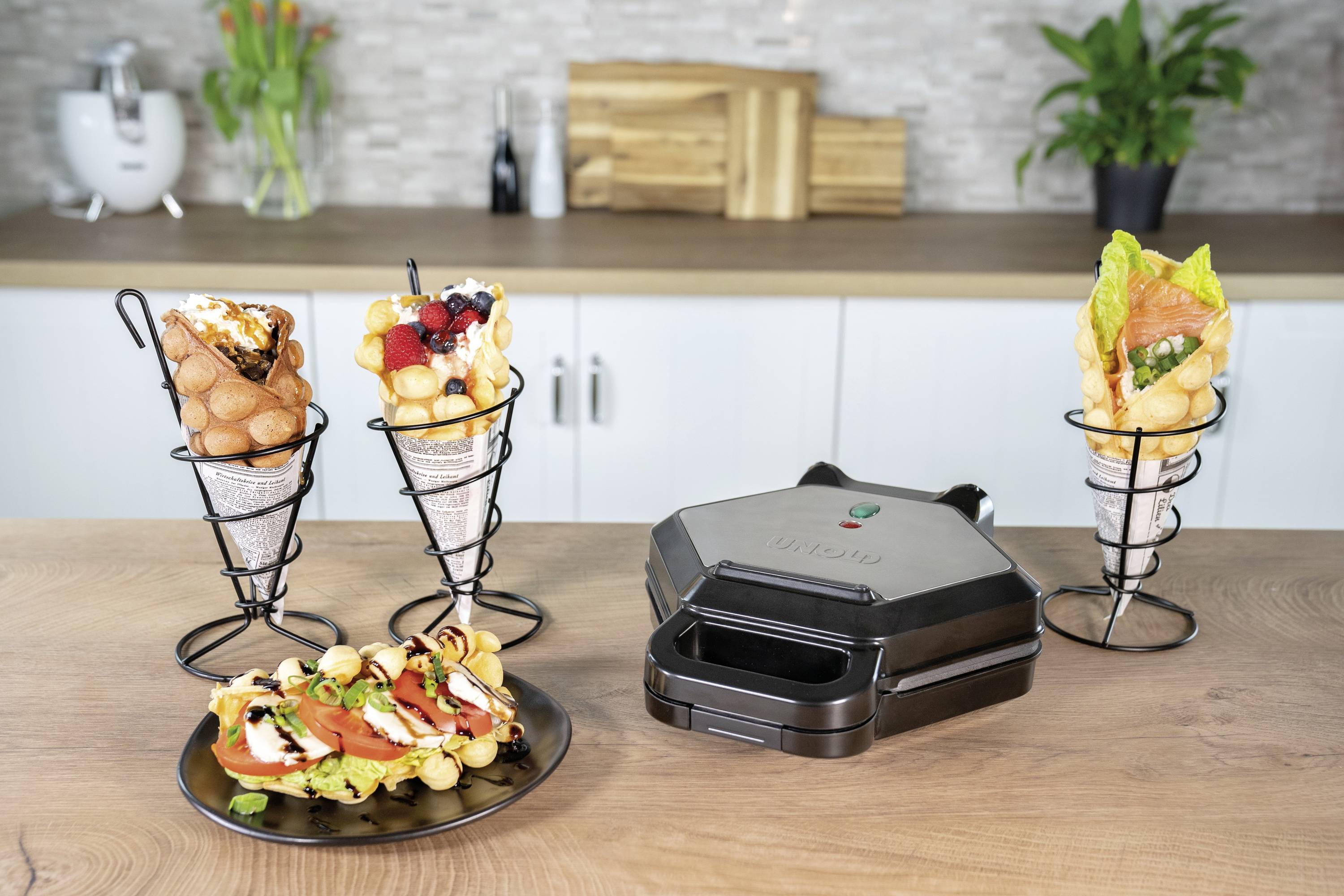 Un elettrodomestico da cucina è posizionato su un tavolo, affiancato da coni di cialda riempiti con frutta e panna, insieme a una prelibatezza di waffle salata.