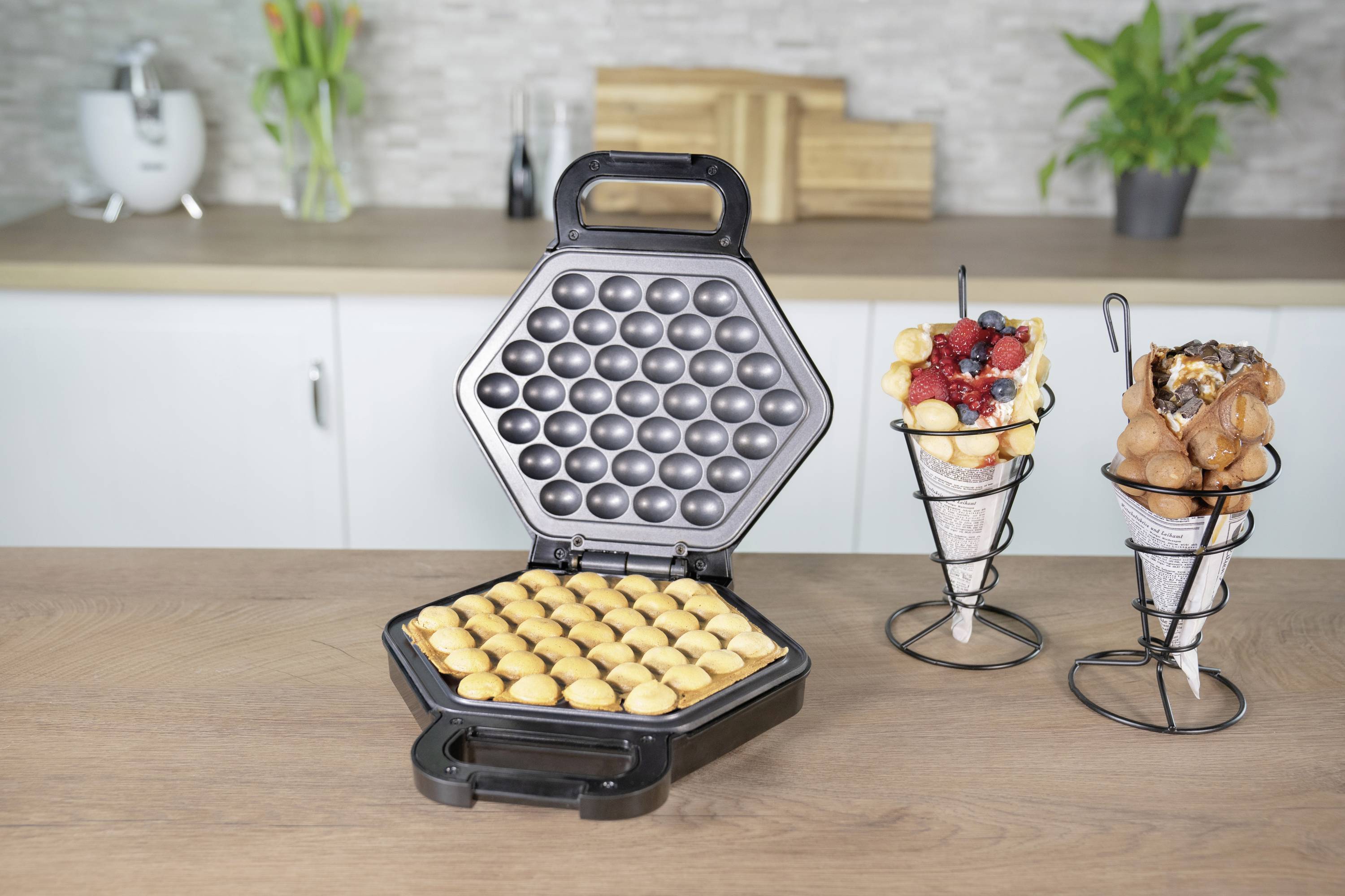 Un ferro per waffle aperto con motivo a nido d'ape rotondo su un tavolo da cucina; accanto due cialde gelato con palline di waffle ripiene.
