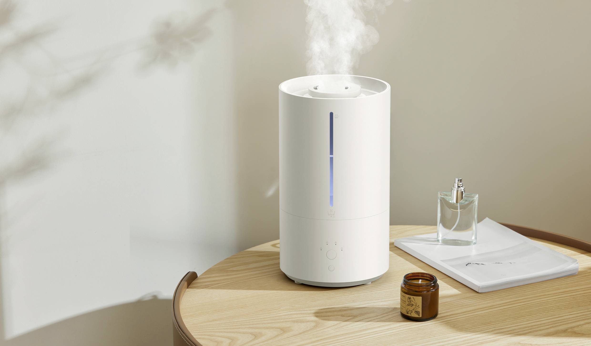 Un umidificatore bianco su un tavolo di legno in una stanza soleggiata. Il vapore sale dal dispositivo. Accanto, una candela profumata e una bottiglia di vetro.