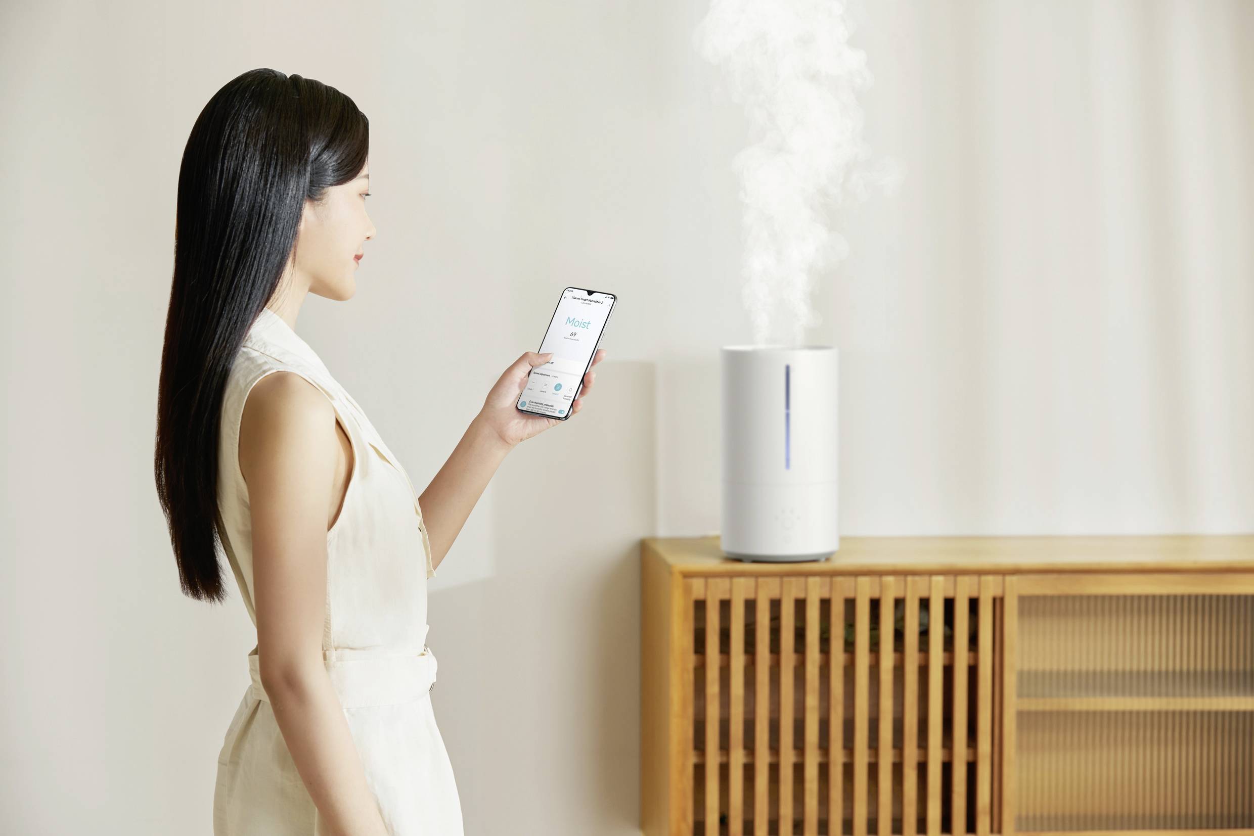 Una donna controlla un umidificatore tramite il suo smartphone. L'umidificatore è posizionato su un tavolo e emette vapore visibile.