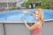 Una donna inserisce una pasticca per la disinfezione della piscina in una piscina fuori terra blu. Sullo sfondo è visibile un giardino.