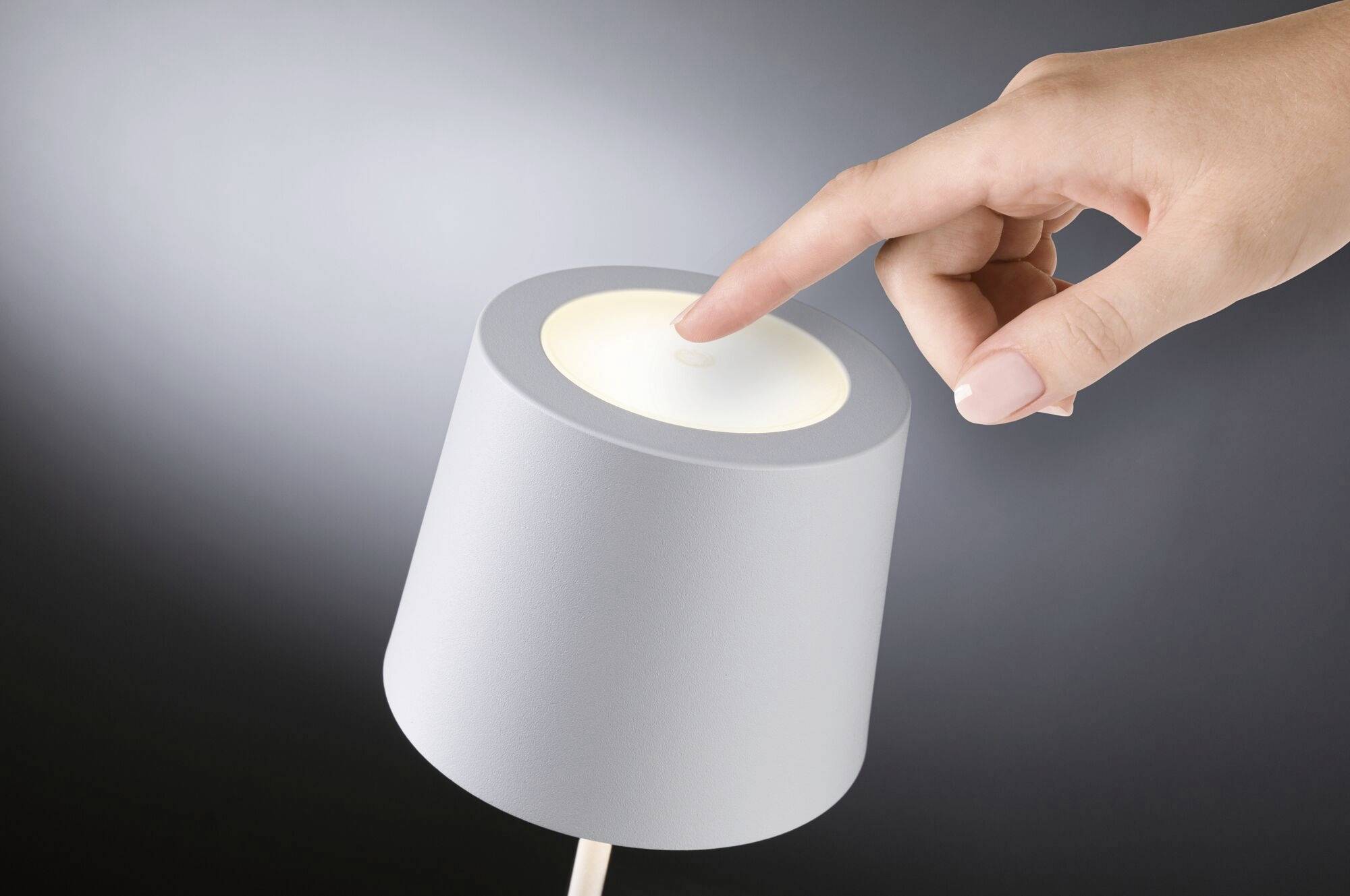 Una mano sfiora delicatamente una lampada da tavolo rotonda e luminosa, sullo sfondo di un ambiente buio.