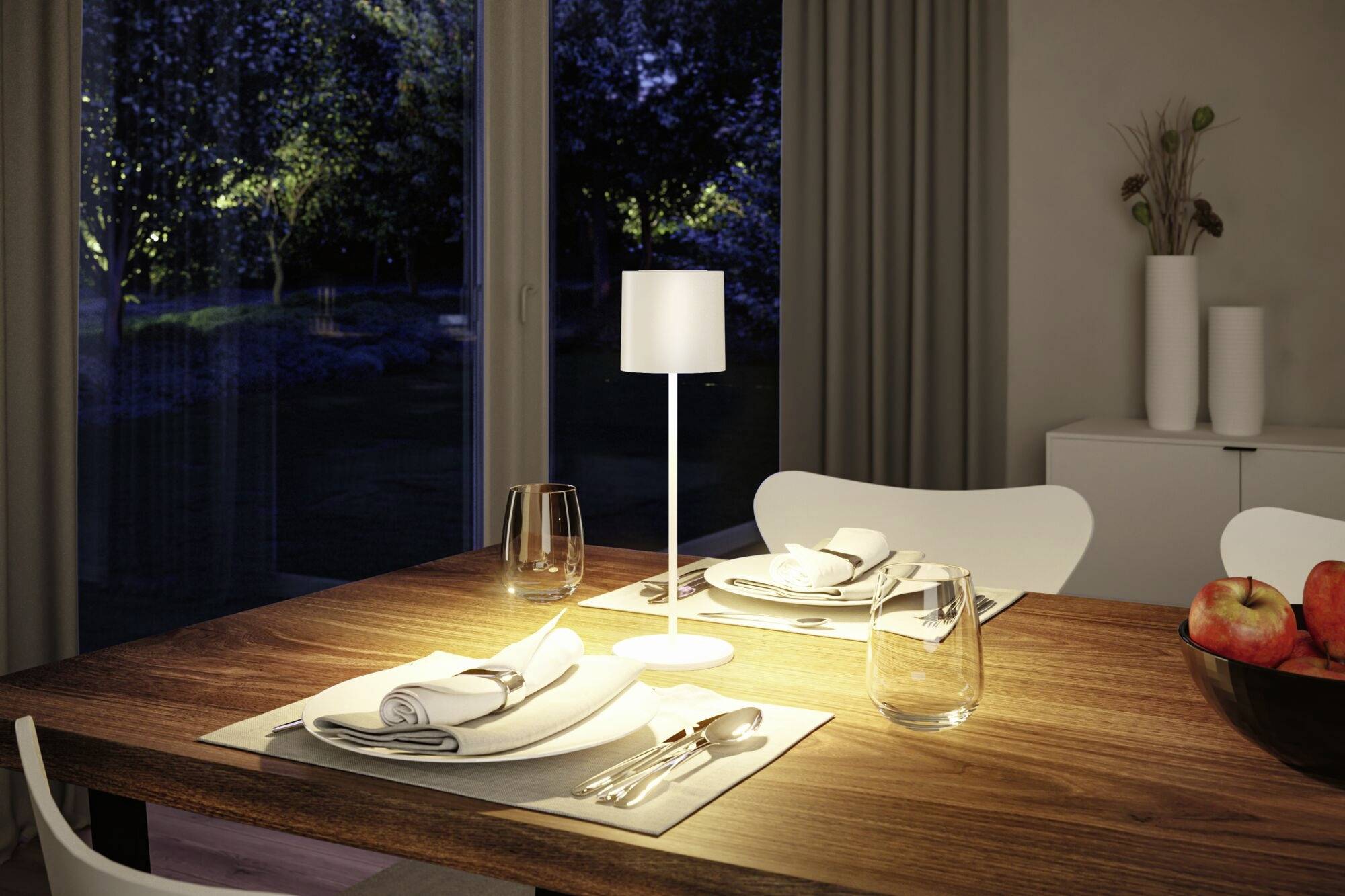 Un tavolo da pranzo elegantemente apparecchiato con un runner bianco, stoviglie e posate. Una lampada illumina il tavolo. Sullo sfondo, finestre con una vista serale.