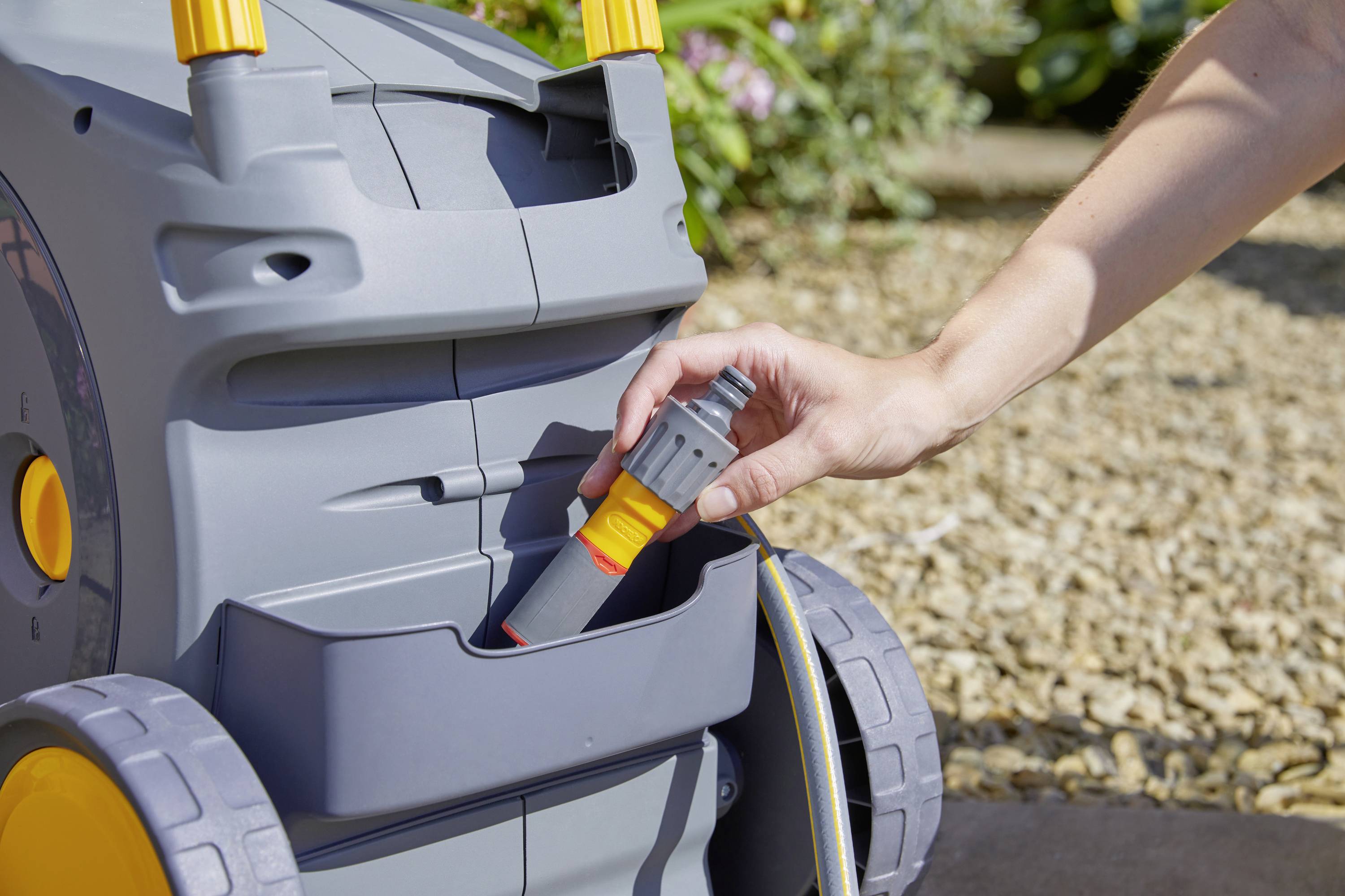 Una mano inserisce un attacco grigio del tubo nel supporto di un carrello porta tubi grigio. Ciottoli e piante sullo sfondo.