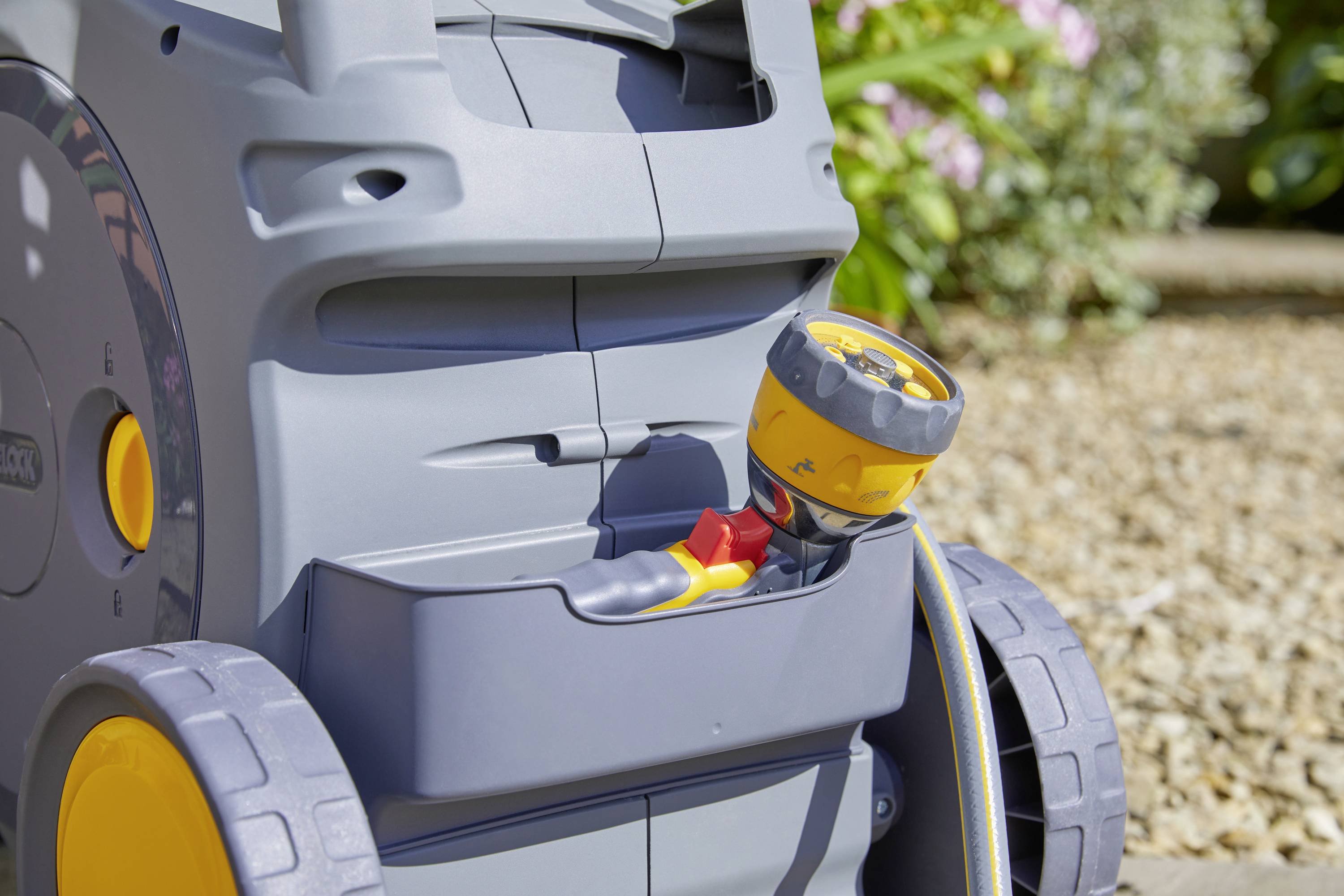 Carrello per tubo da giardino con ugello giallo e ruote grandi sotto la luce del sole su un terreno di ghiaia.