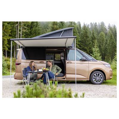 Due persone sono sedute a un tavolo da campeggio accanto a un camper beige con una tenda da sole, parcheggiato in un campeggio nel bosco.