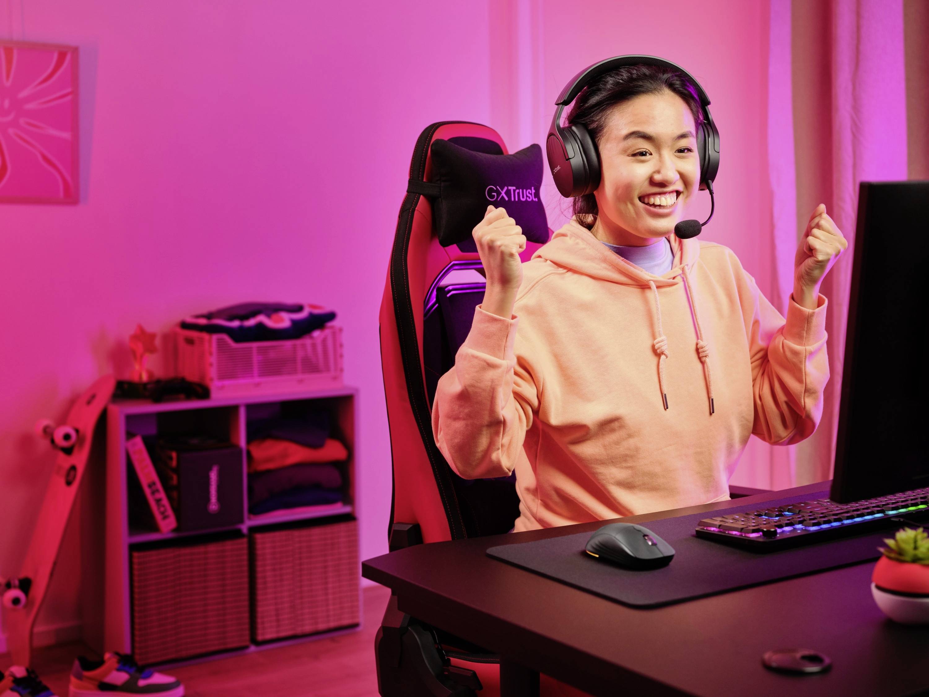 Una persona è seduta davanti a un computer in una stanza illuminata di rosa, indossa un headset ed esulta con i pugni alzati.