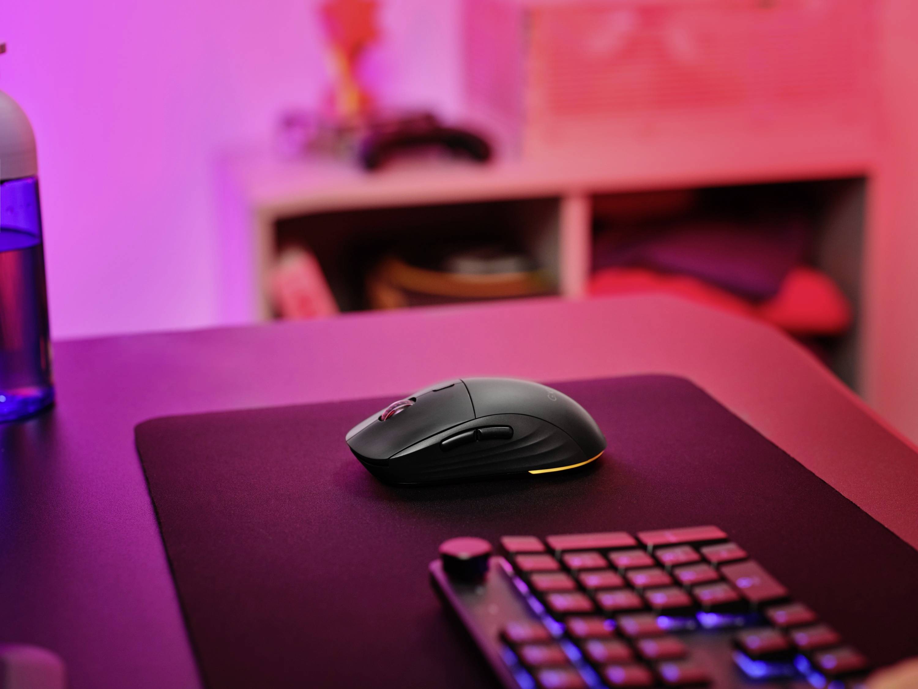 Un mouse wireless su un tappetino per mouse nero, sullo sfondo uno scaffale con oggetti sfocati. La stanza immersa in una luce rosa.