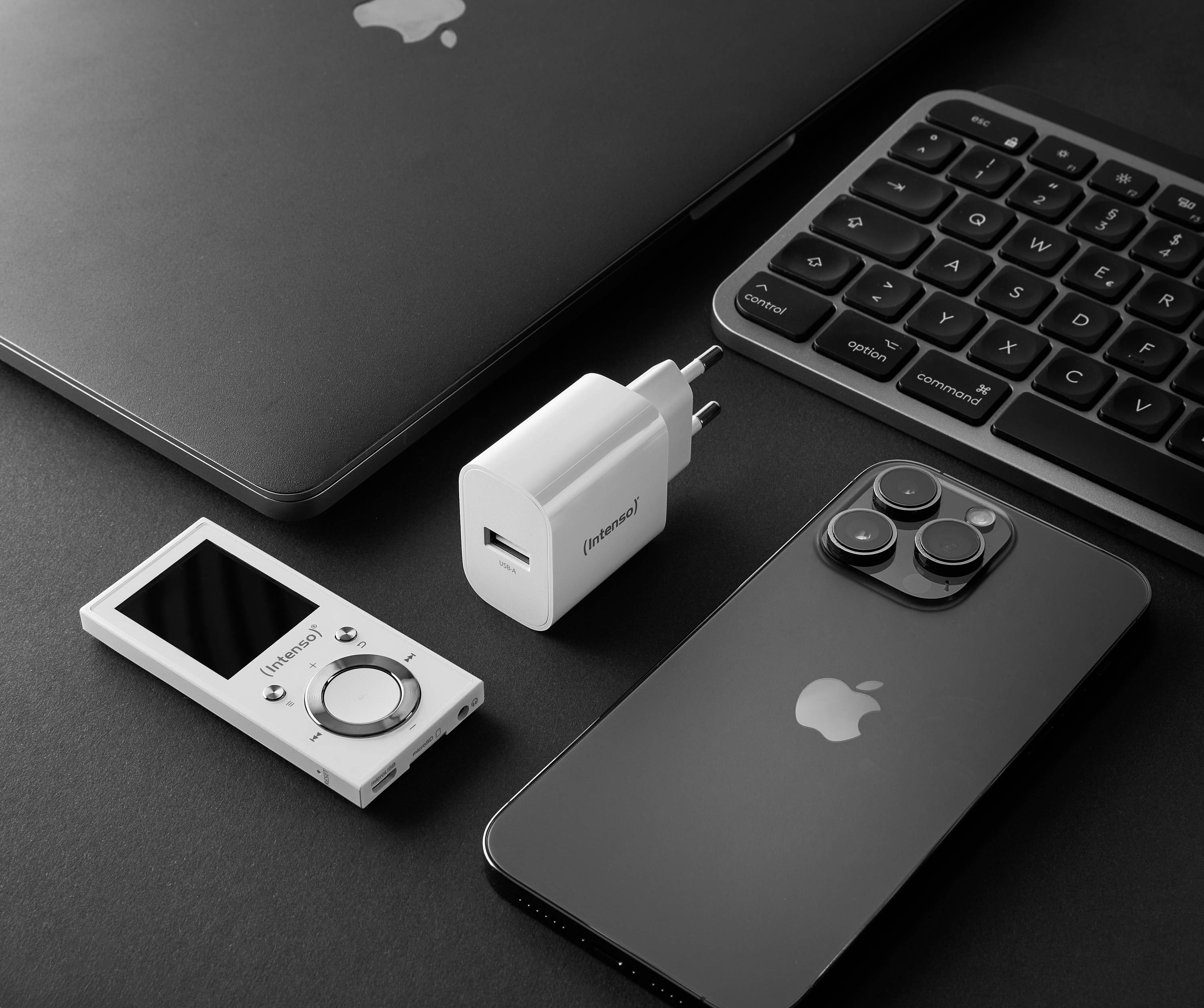 Laptop, smartphone, lettore musicale portatile e caricabatterie sono posati su un tavolo. Tutti i dispositivi sono in bianco e nero.